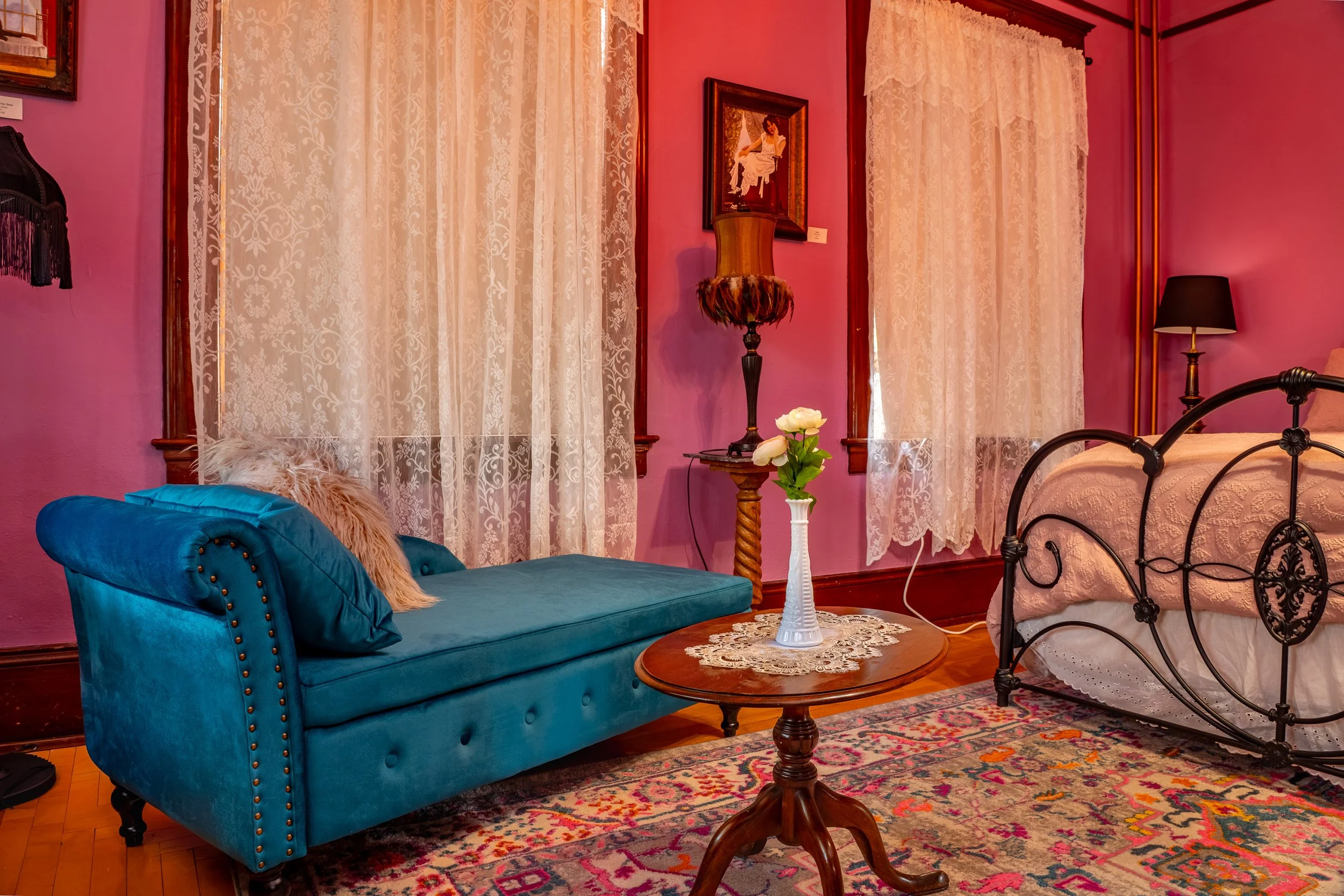 A cozy bedroom with pink walls and lace curtains. There is a blue velvet tufted sofa with a pillow, a wooden round coffee table with a vase of flowers, a brass bed frame, and a nightstand with a black table lamp. The wall features a framed painting of a woman sitting, and there is a tall floor lamp beside the bed.