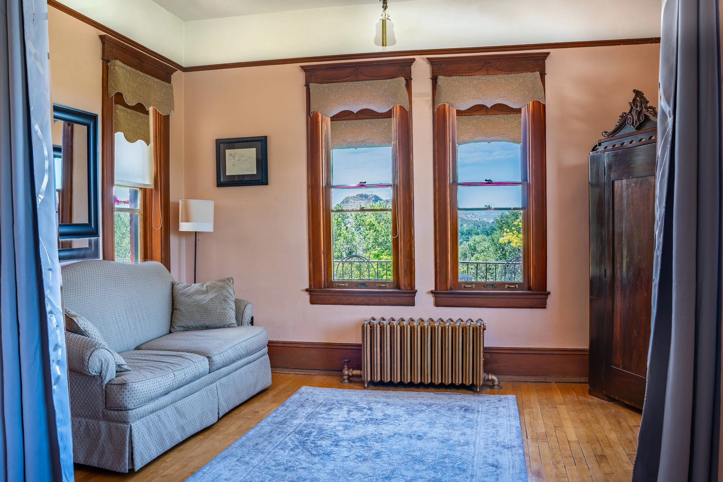 Living room with a beige sofa, a white lamp, framed artwork, three tall windows with wooden frames and beige curtains, an old radiator, hardwood floors, and a view of trees and mountains outside the windows.
