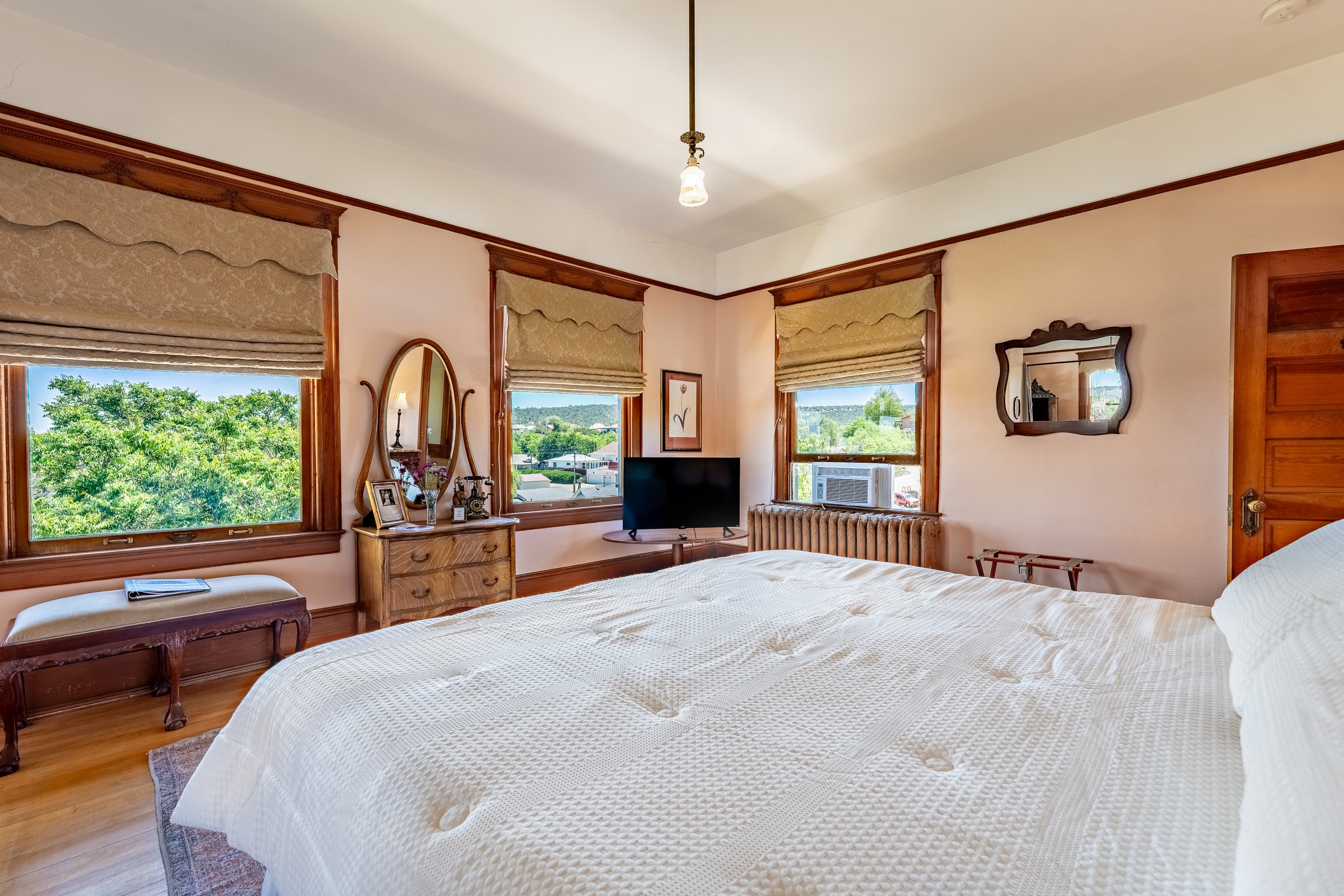 A cozy bedroom with a large bed covered in a white textured bedspread, three windows with beige roman shades, and wooden trim. The room features vintage furniture, including a small desk with a mirror, a television, a radio, and framed pictures. Green trees and a blue sky are visible outside the windows.