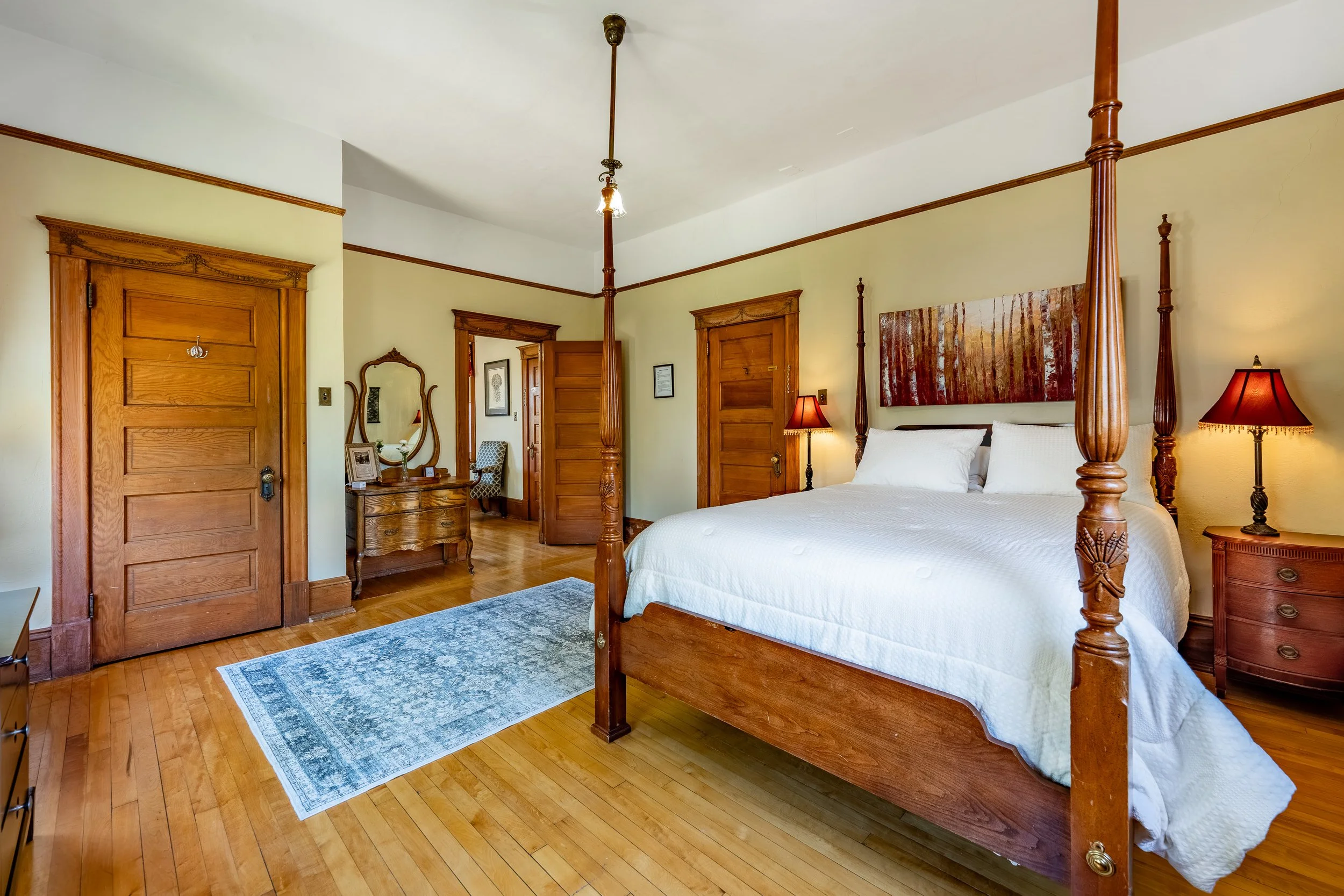 A bedroom with a four-poster wooden bed, two red lamps, a wooden dresser with a mirror, and wooden doors and trim.