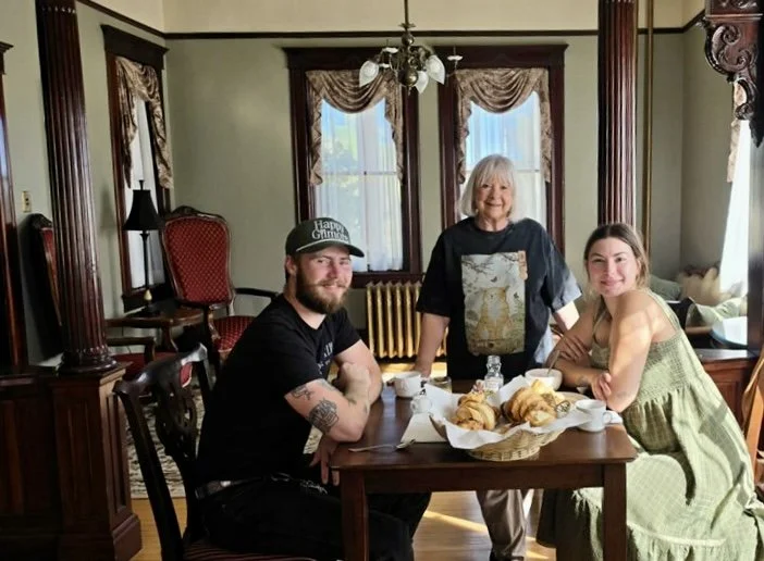 Three people sitting and standing around a wooden dining table with breakfast food in a vintage-style dining room with ornate wood trim, curtains, and a chandelier.