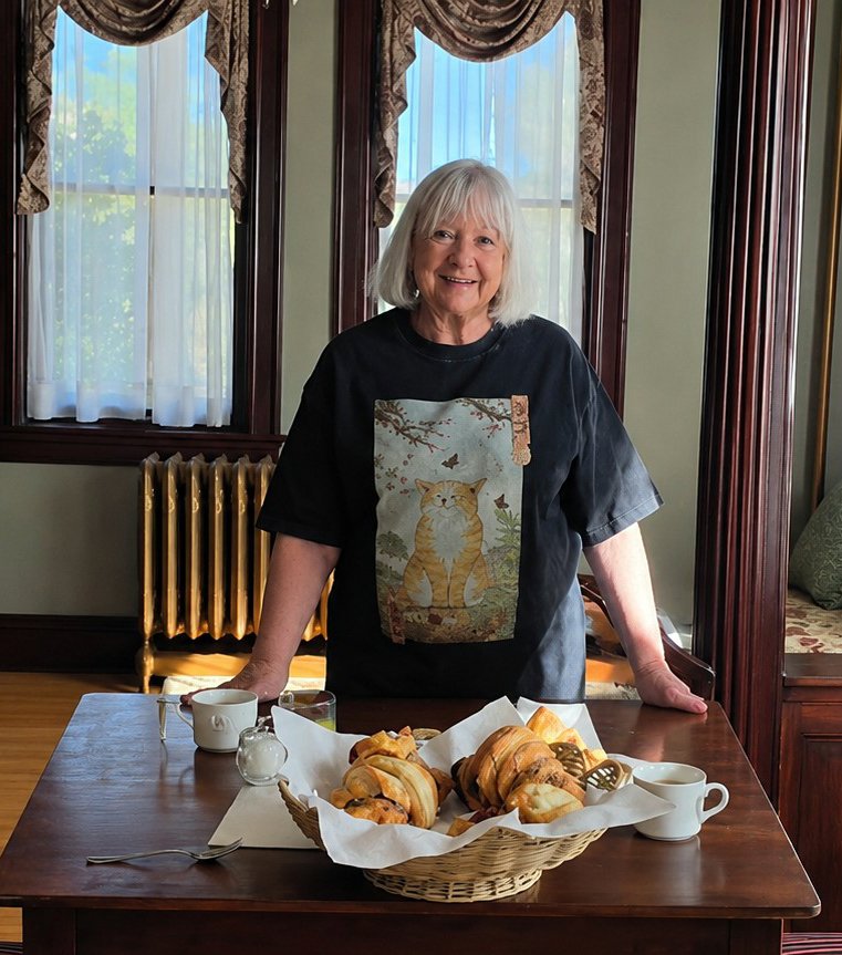 Jeanne, owner of Tarabino Inn  standing behind a wooden table with a basket of croissants, cups, and a spoon, in a vintage dining room with large windows, curtains, and wooden trim.