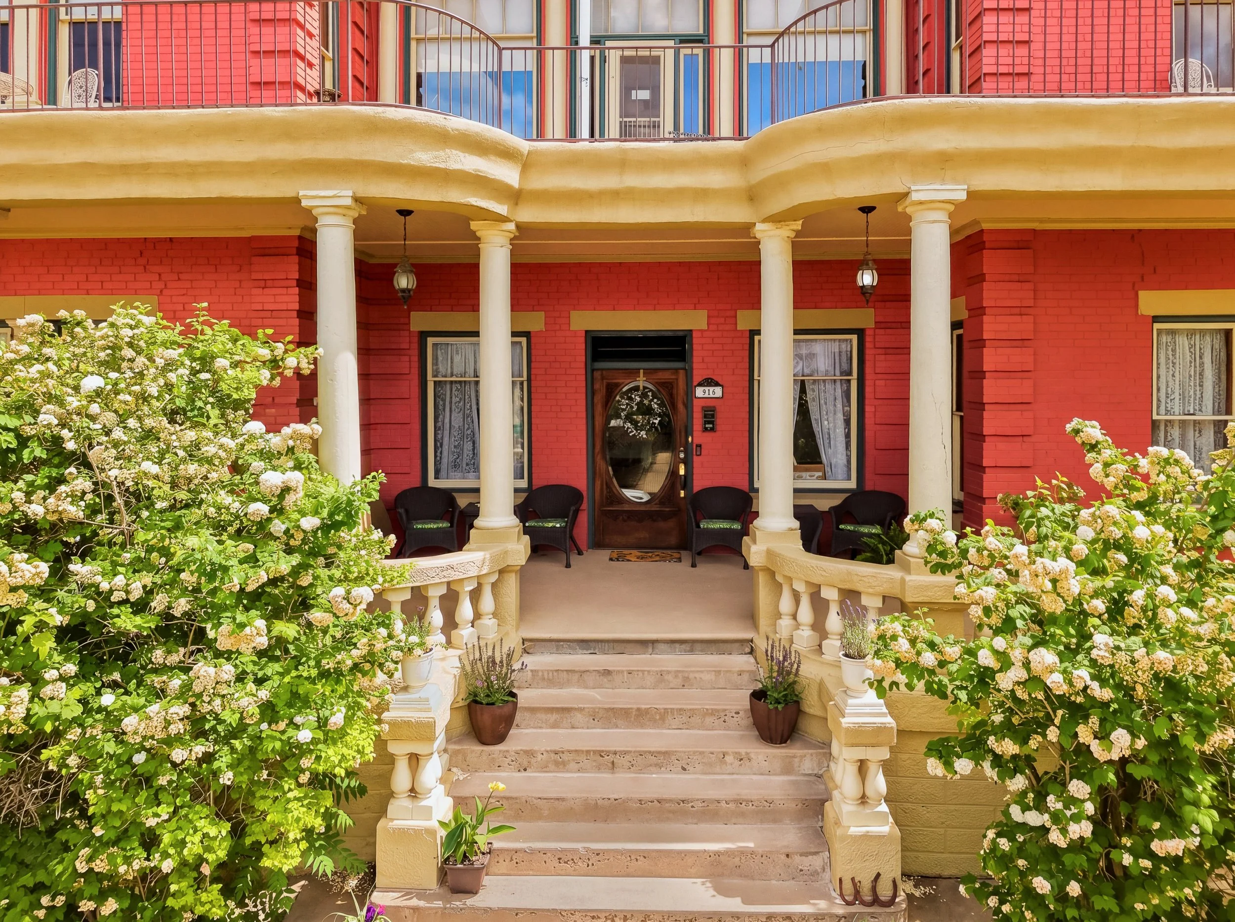 Front porch of a red brick house with white columns, potted plants, two entrance doors, and greenery with white flowers in front.