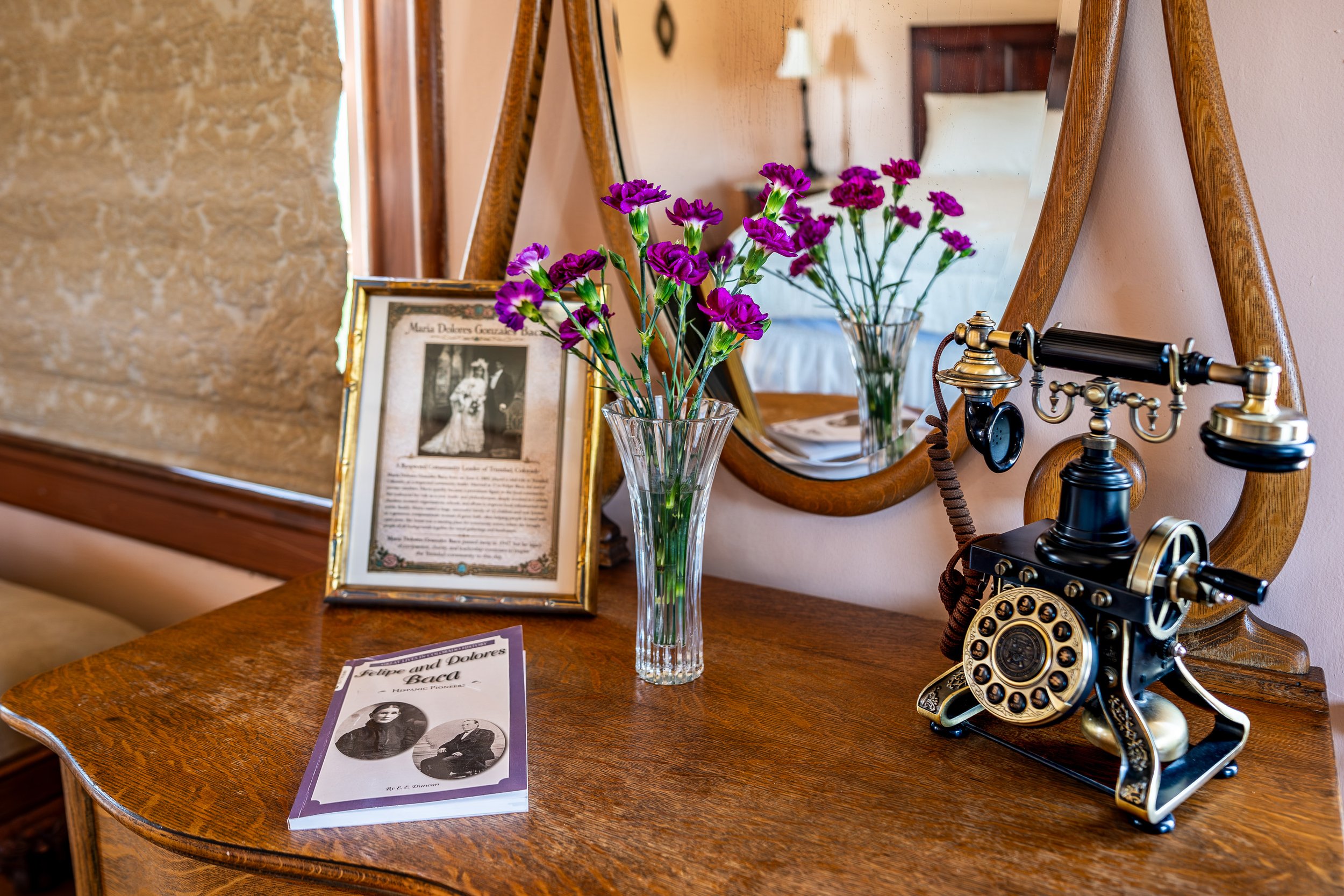A wooden table with a framed black-and-white photograph, a purple flower bouquet in a glass vase, and an antique black and brass telephone. In the background, there's a mirror reflecting part of the room.