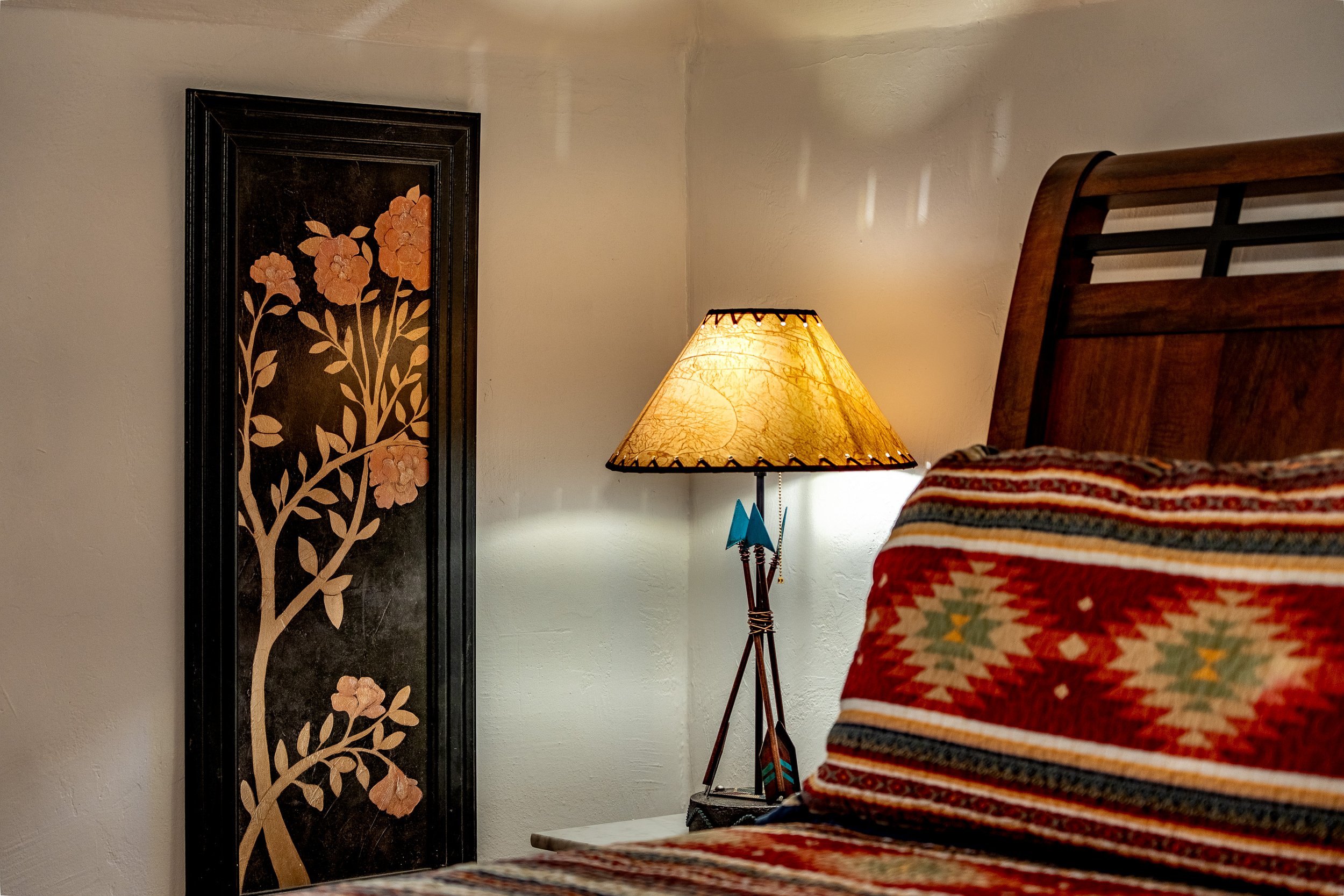 A bedroom corner with a floral wall hanging, a lit table lamp, and a bed with a multicolored striped blanket.