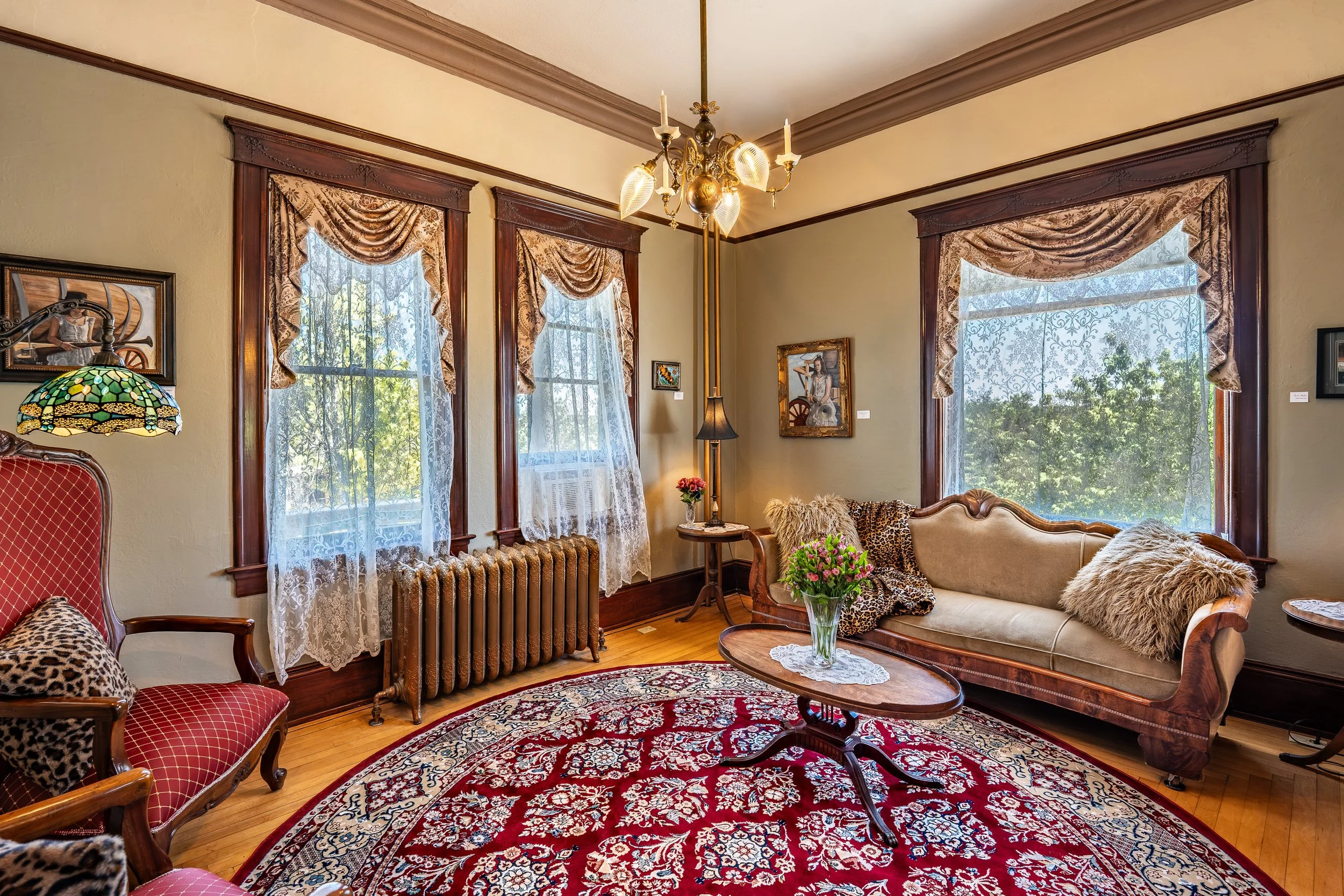 A vintage living room with large windows, lace curtains, a chandelier, a rug, a sofa with fur pillows, and antique furniture and artwork.