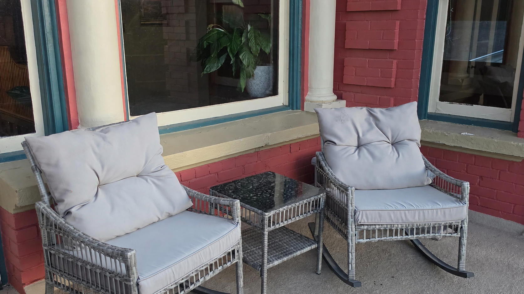 A cozy outdoor seating area with two rocking chairs that have white cushions, a small black table with a marble top, set against a brick and windowed wall with a potted plant visible through the window.