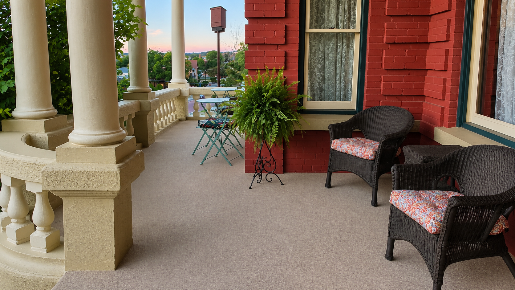 Balcony with cream-colored columns, potted fern, red brick wall, wicker chairs with floral cushions, and outdoor table with chairs, overlooking a neighborhood at sunset.