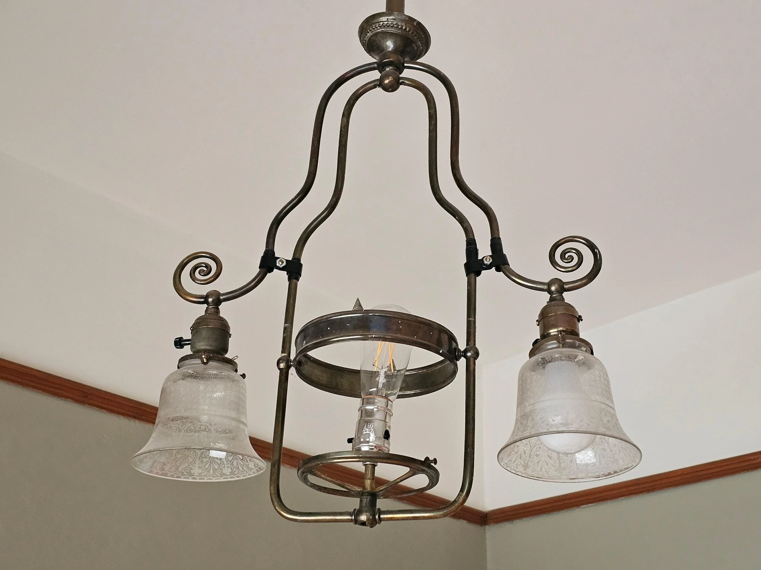 A vintage chandelier with four frosted glass lampshades hanging from a metal frame, mounted on a ceiling with crown molding.