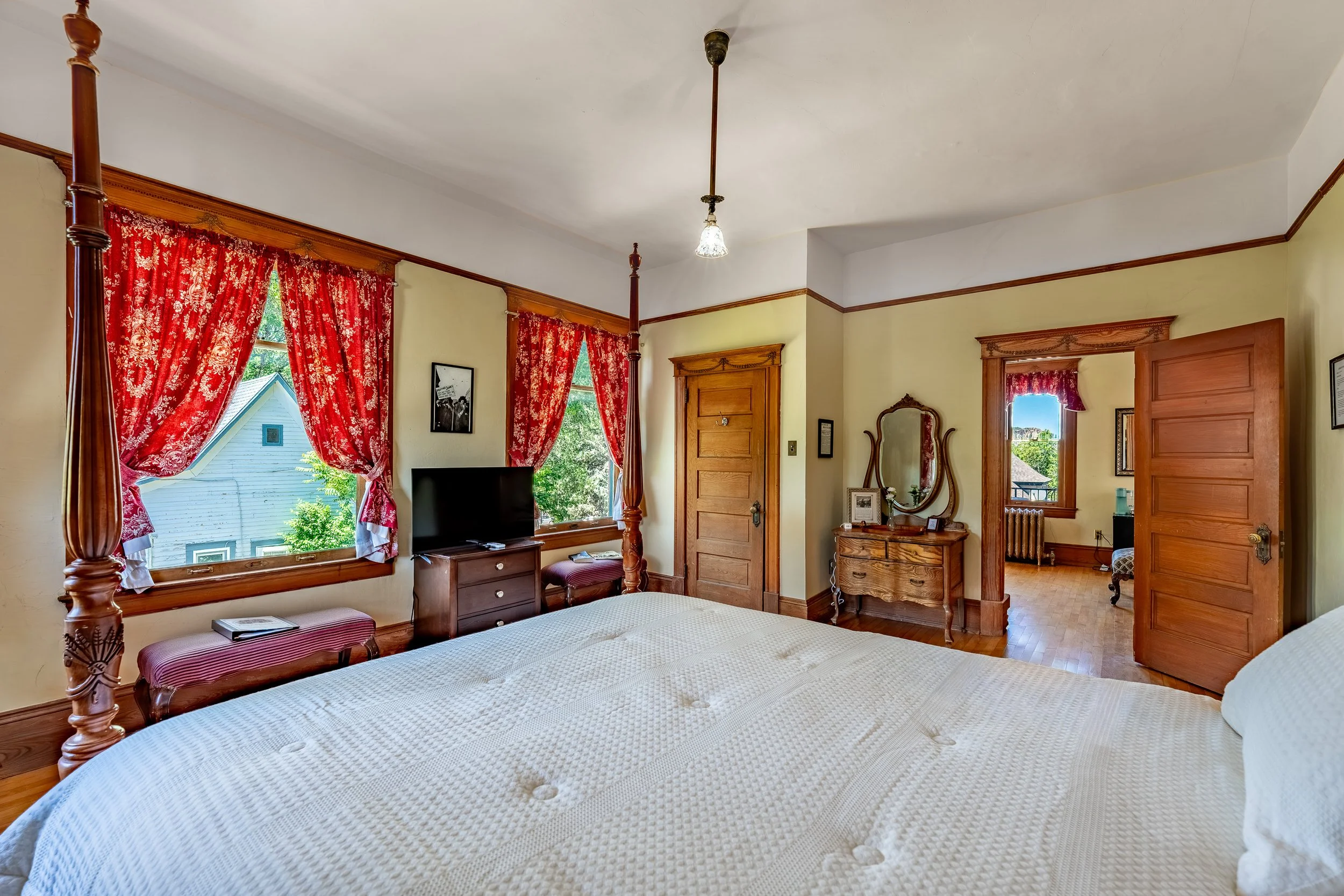 A bedroom with a four-poster bed, cream-colored quilt, large windows with red floral curtains, a small dresser with a TV, a mirror, and some framed pictures on the walls. Wooden trim and doors add a classic touch.