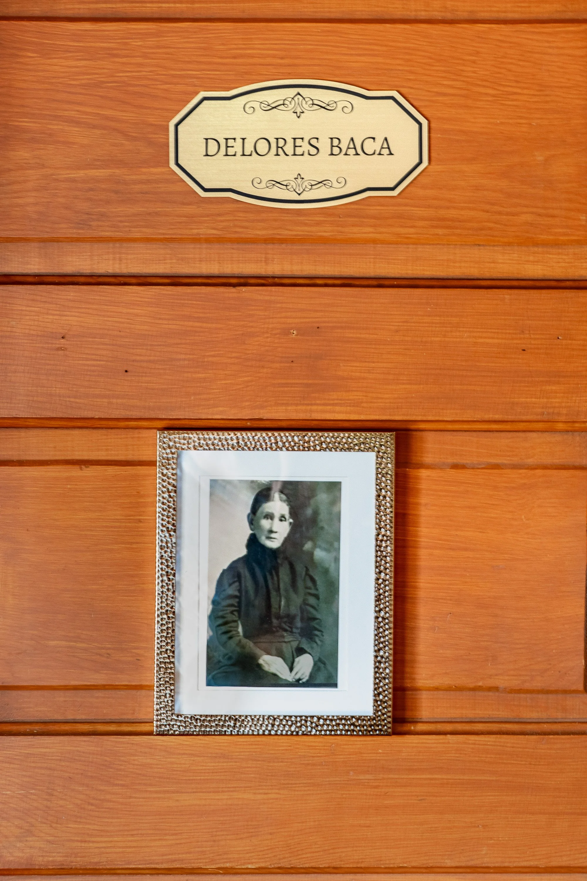 A framed black-and-white photograph of a woman in a dark jacket, sitting with her hands resting on her lap, mounted on a wooden wall.