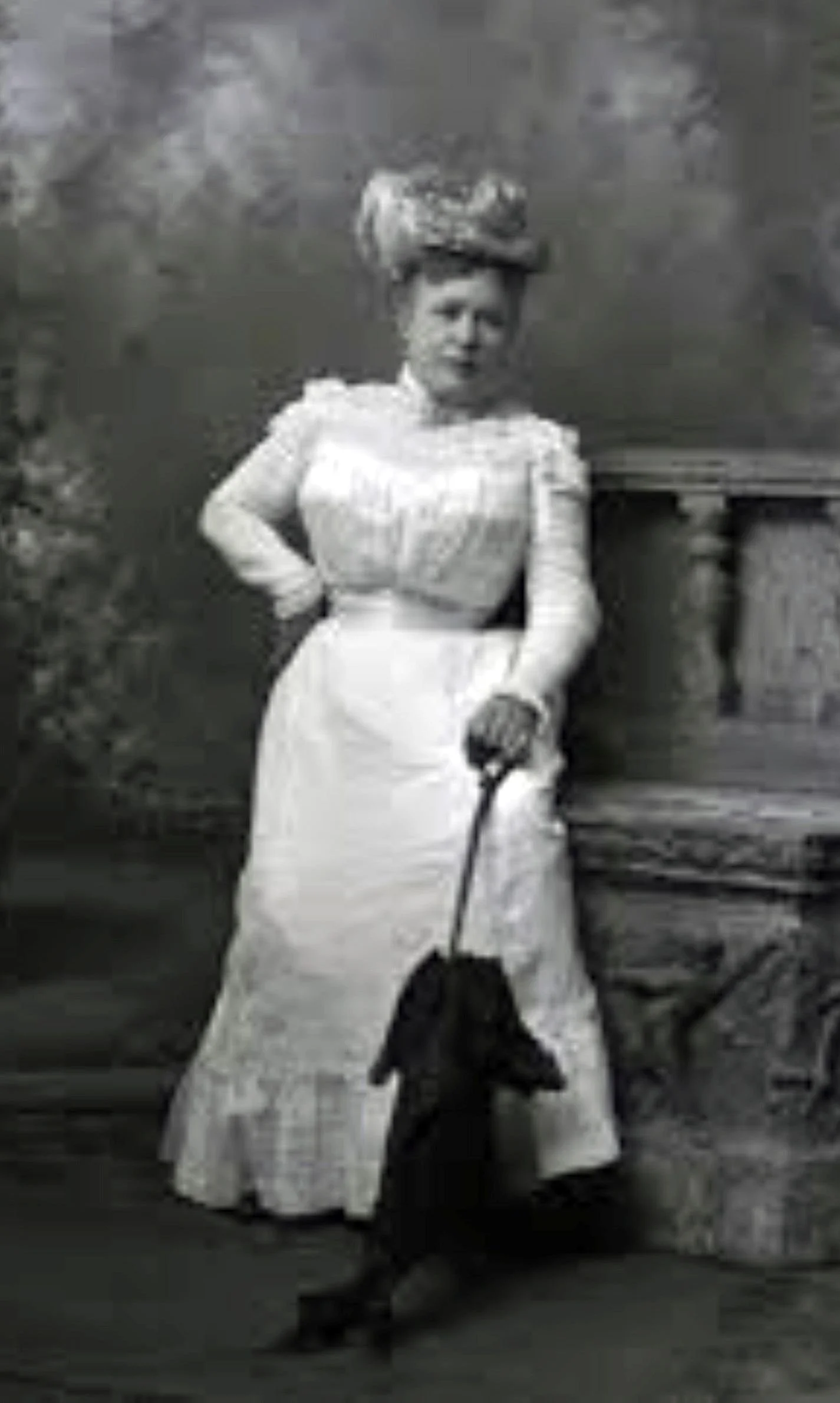 A woman in a long skirt and blouse standing outdoors, with a hat on her head. She is holding an umbrella or parasol and looking at the camera. There is a dog in front of her, sitting and facing the camera. The background features a rustic scene with trees and a wooden bench.
