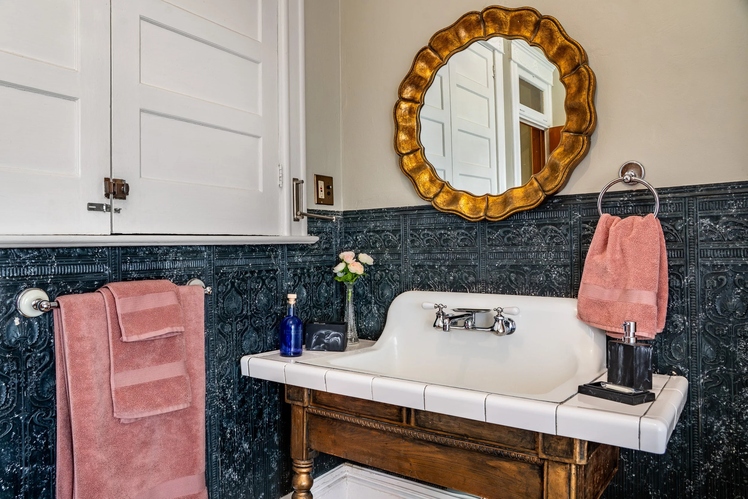 A vintage bathroom with a white cast iron bathtub, a large ornate gold mirror, pink towels hanging on a chrome towel bar, a small vase with pink roses, blue bottles, and a dark wood vanity with a white tiled top. The bathroom features dark, decorative tile on the lower half of the wall, a beige upper wall, and a white door.