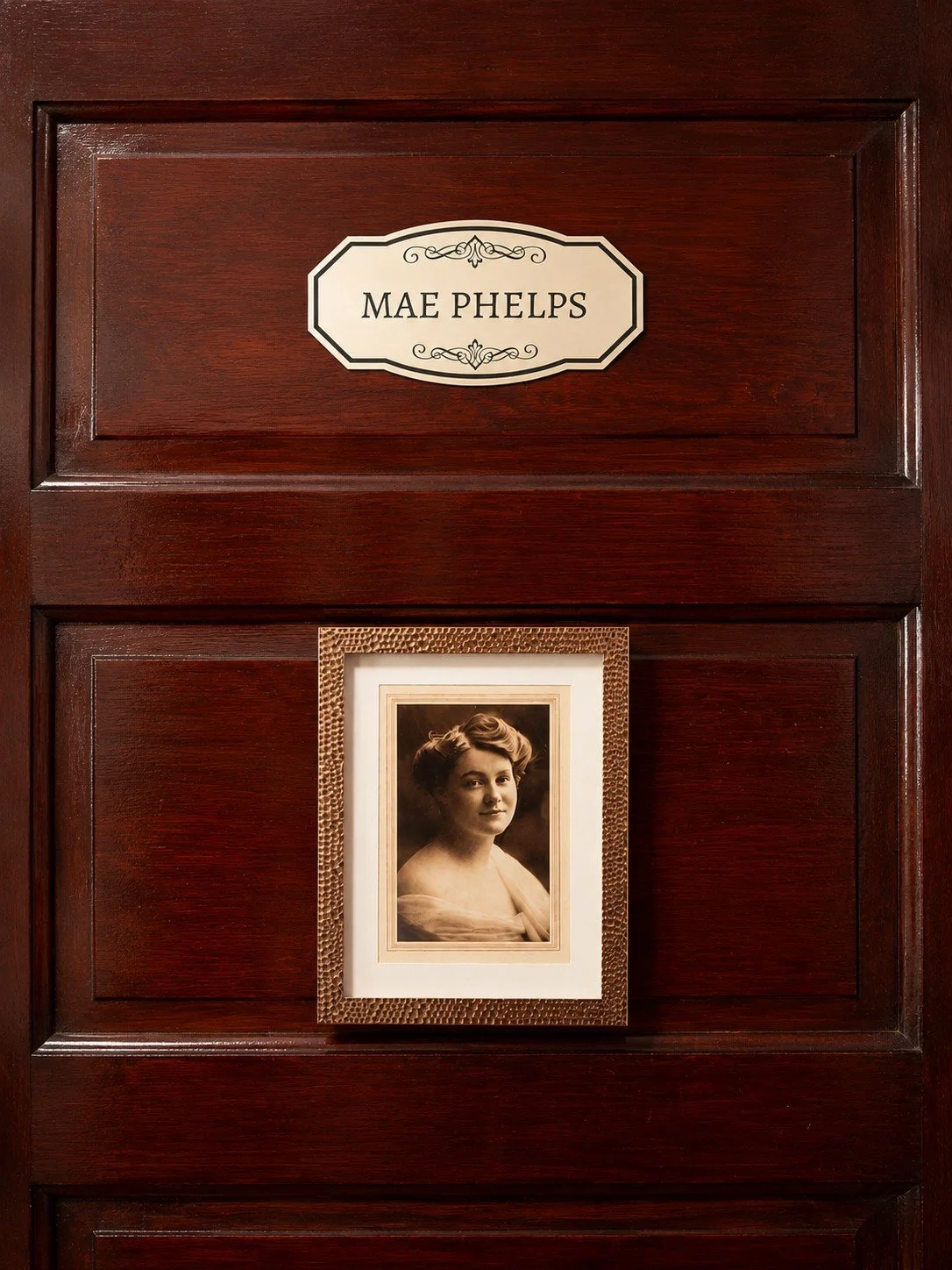 Close-up of a wooden door with a nameplate reading 'Mae Phelps' and a sepia portrait photograph of a woman framed and hung below the nameplate.