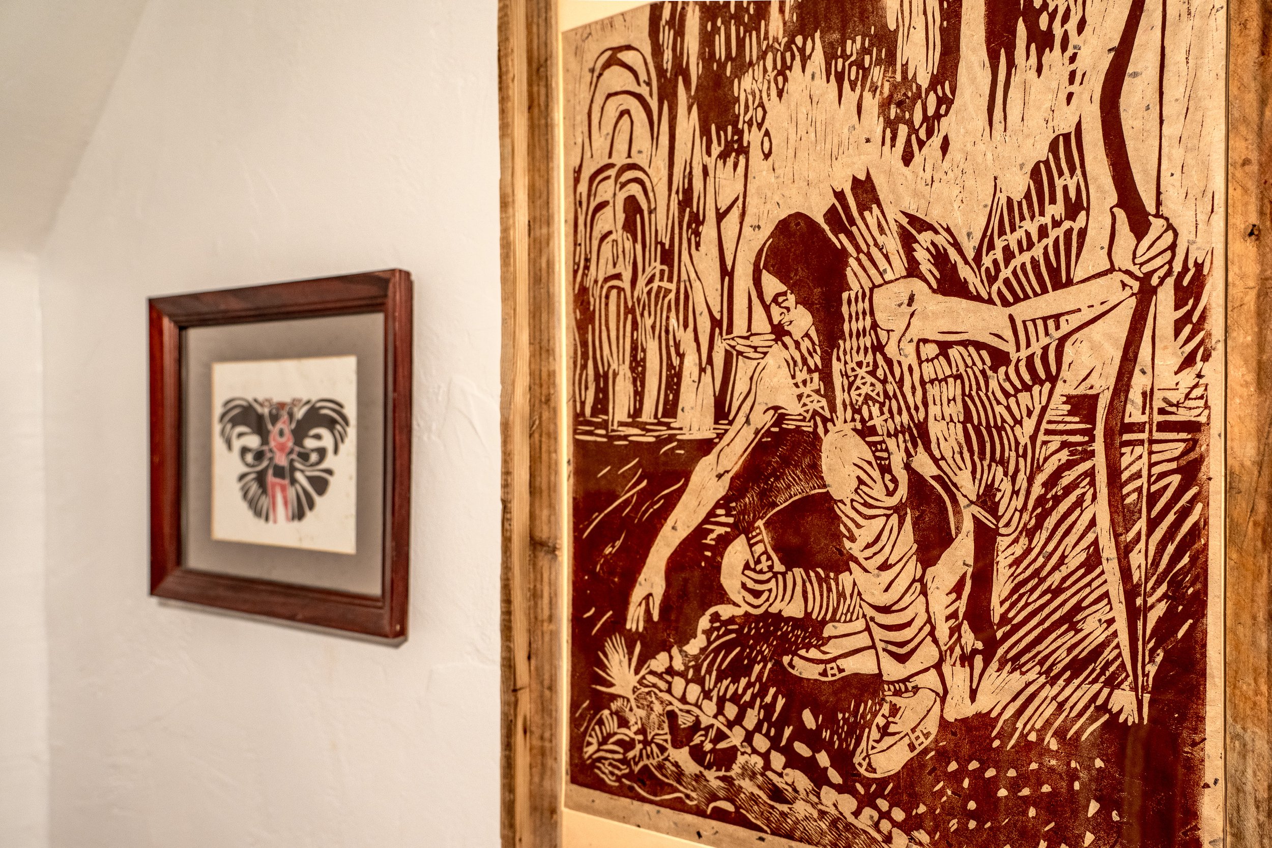 Artwork in a wooden frame depicting stylized female figures in a natural setting, with trees and water, in earthy tones.