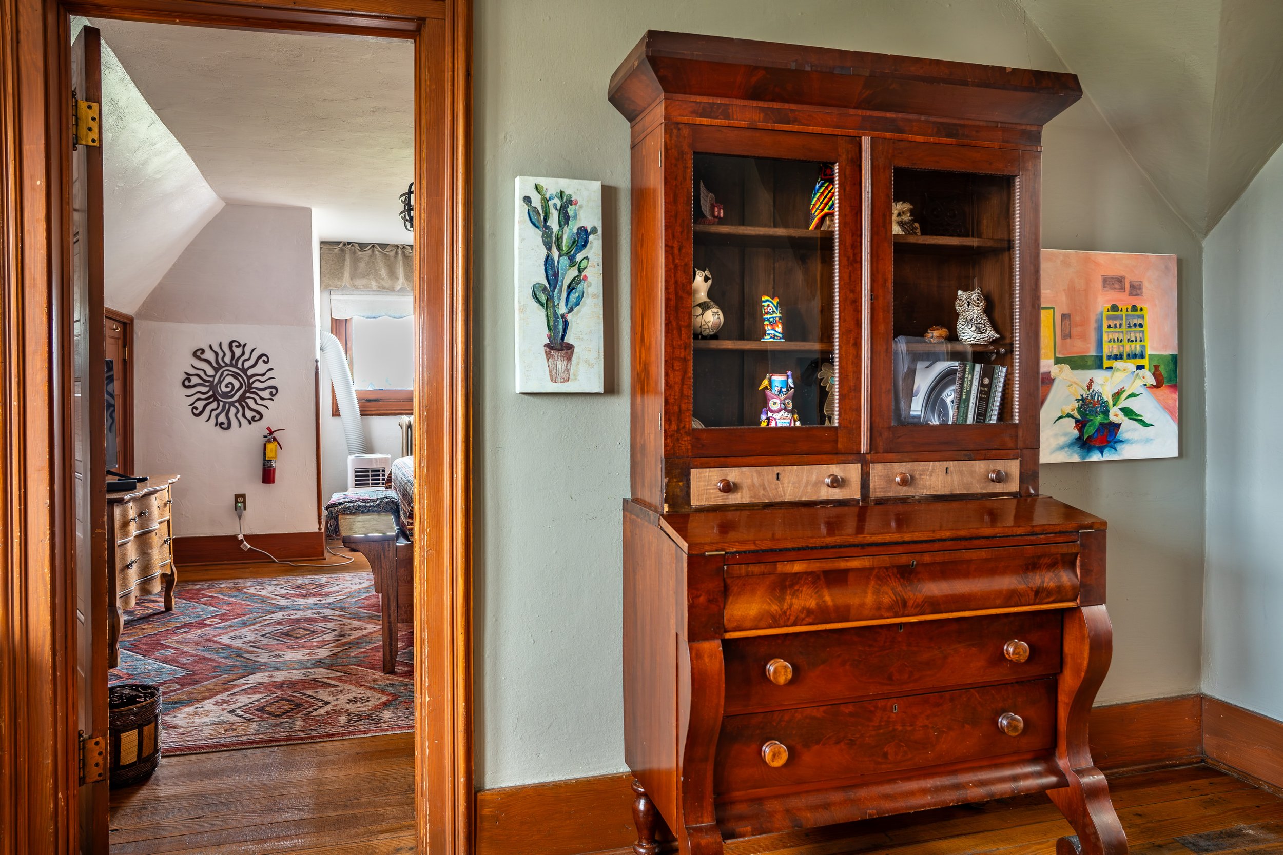 A wooden roll-top desk with multiple drawers stands against a pale green wall, reflecting light. Behind, through a doorway, a room with a colorful patterned rug, a wall decoration resembling a sun or sunflower, and a window with a beige shade can be seen. Artworks and a cactus painting hang on the walls.
