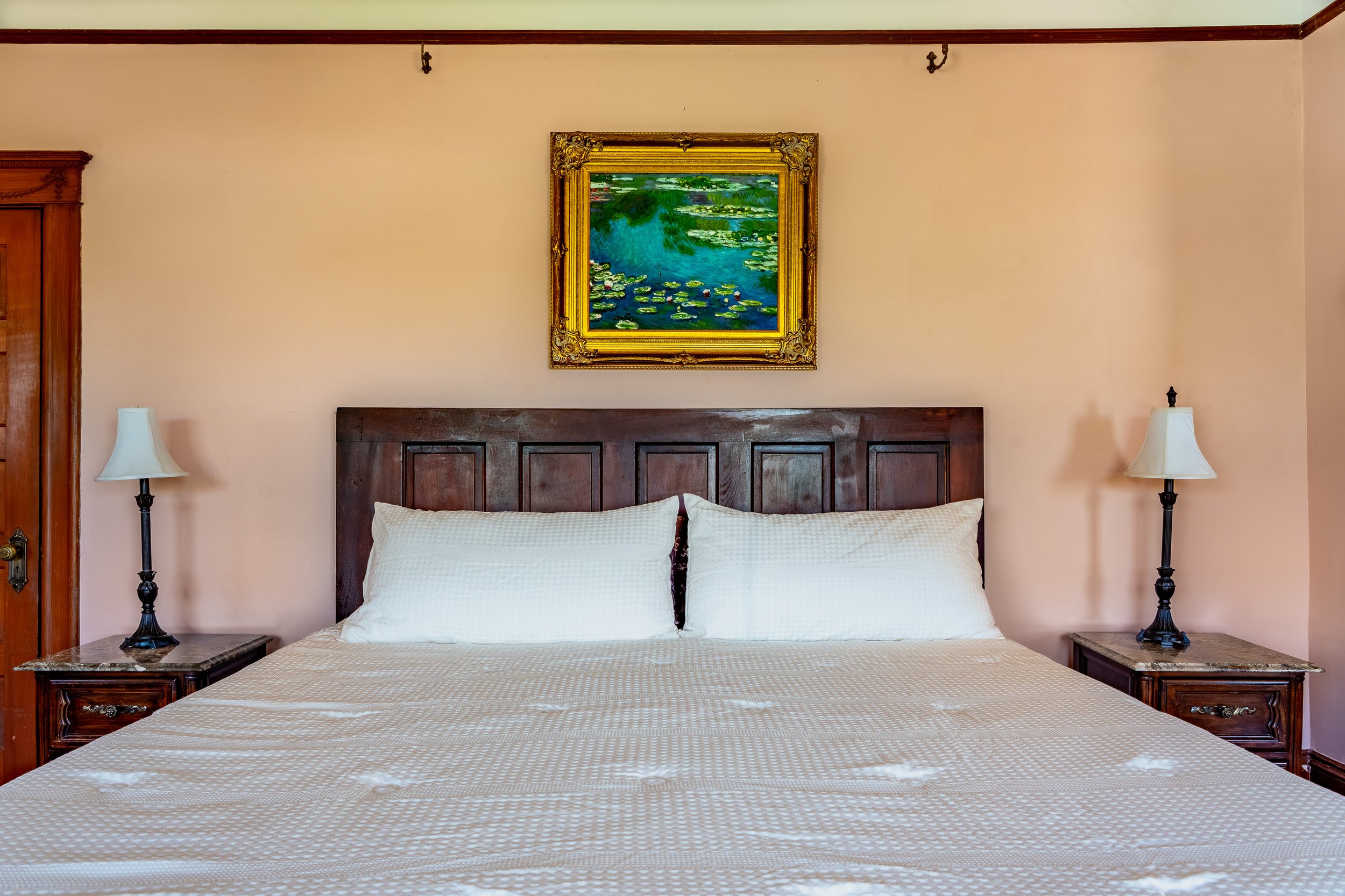 A bedroom with a large bed covered with white bedding, two wooden nightstands, each with a white lamp, a wooden headboard, and a framed painting of water lilies on the wall above the headboard.