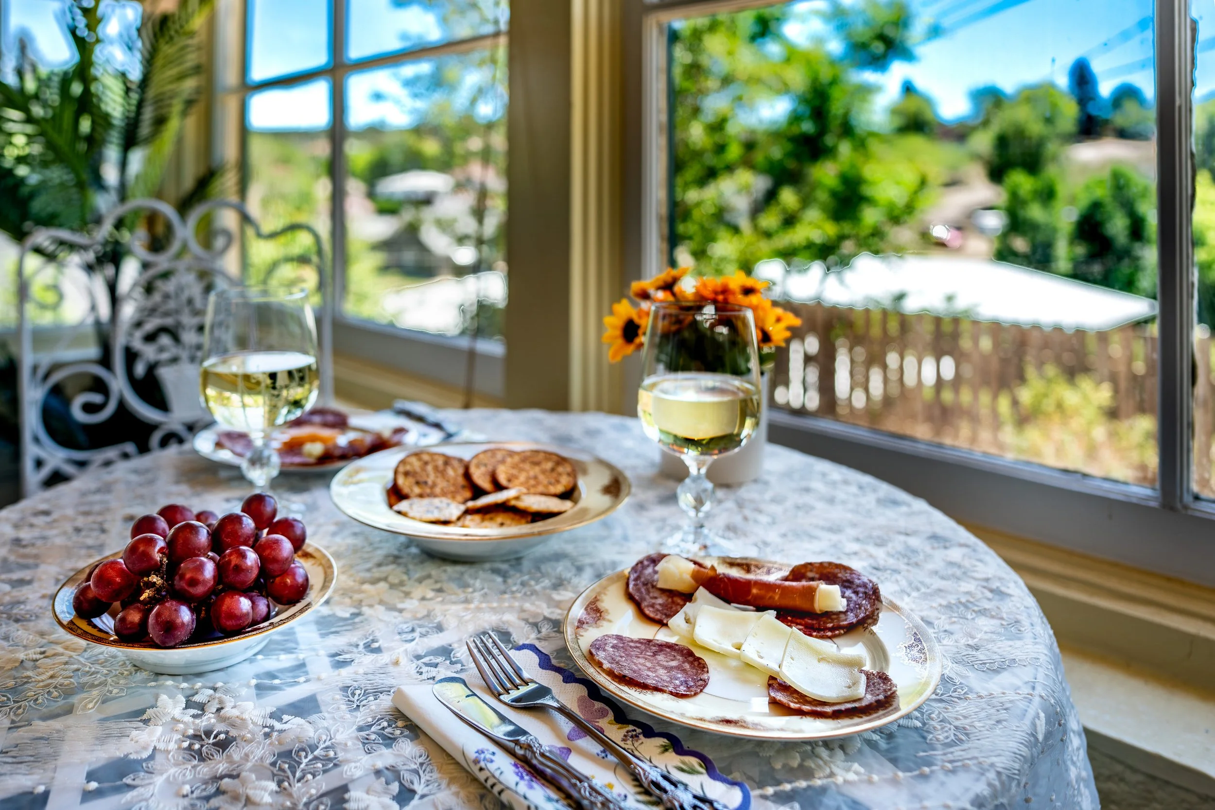 A table set for breakfast with plates of salami and cheese, a bowl of red grapes, and glasses of white wine. Sunlight streams through large windows revealing a lush outdoor view.