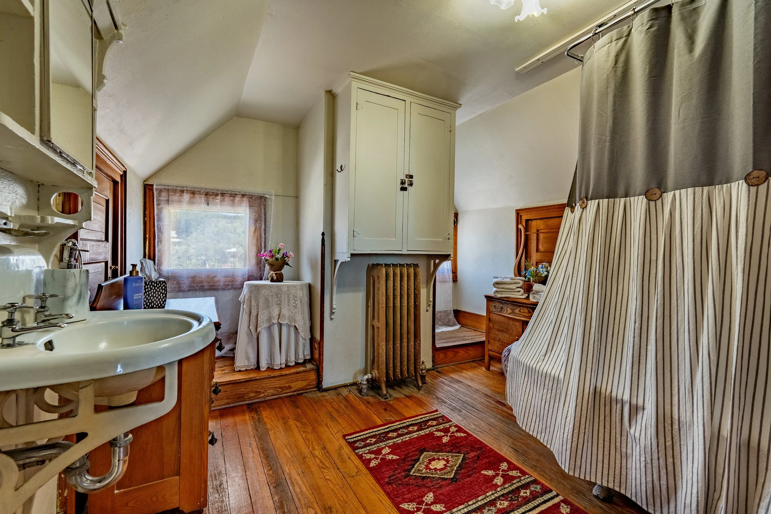 A cozy attic bathroom with a small sink, a mirror, a small side table with a vase of flowers, a window with sheer curtains, an old radiator, a wardrobe, a wooden bed frame, a cloth shower curtain, and a red patterned rug on a wooden floor.