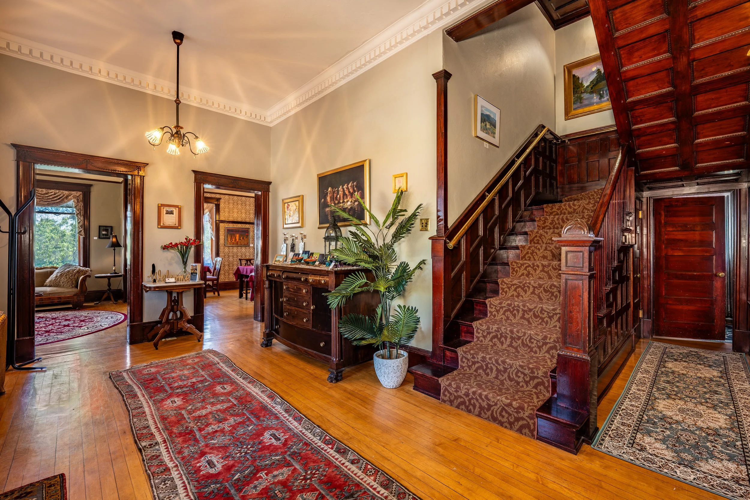 Interior view of a vintage-style house with hardwood floors, area rugs, a staircase with a patterned carpet runner, artwork on the walls, and a partial view of a living room with a sofa and floral curtains.