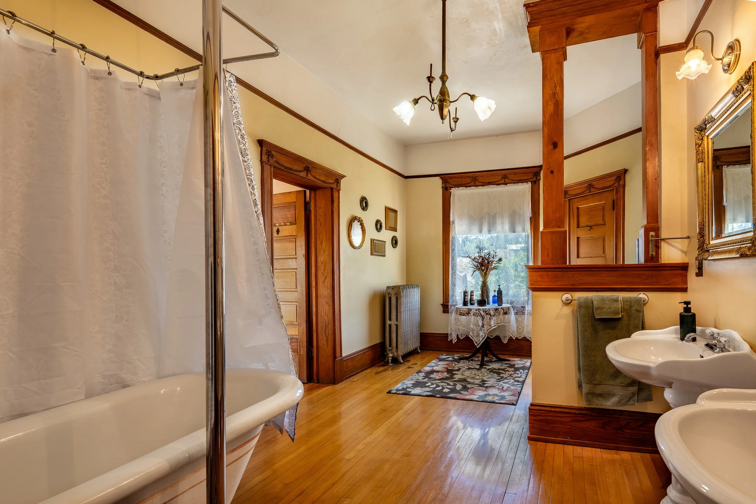 A vintage-style bathroom featuring wood trim and fixtures, a bathtub with a shower curtain, a porcelain sink with a green towel, a mirror with decorative frame, a small round carpet, a window with a lace curtain, and a table with vases, all illuminated by a ceiling chandelier and wall sconce lighting.