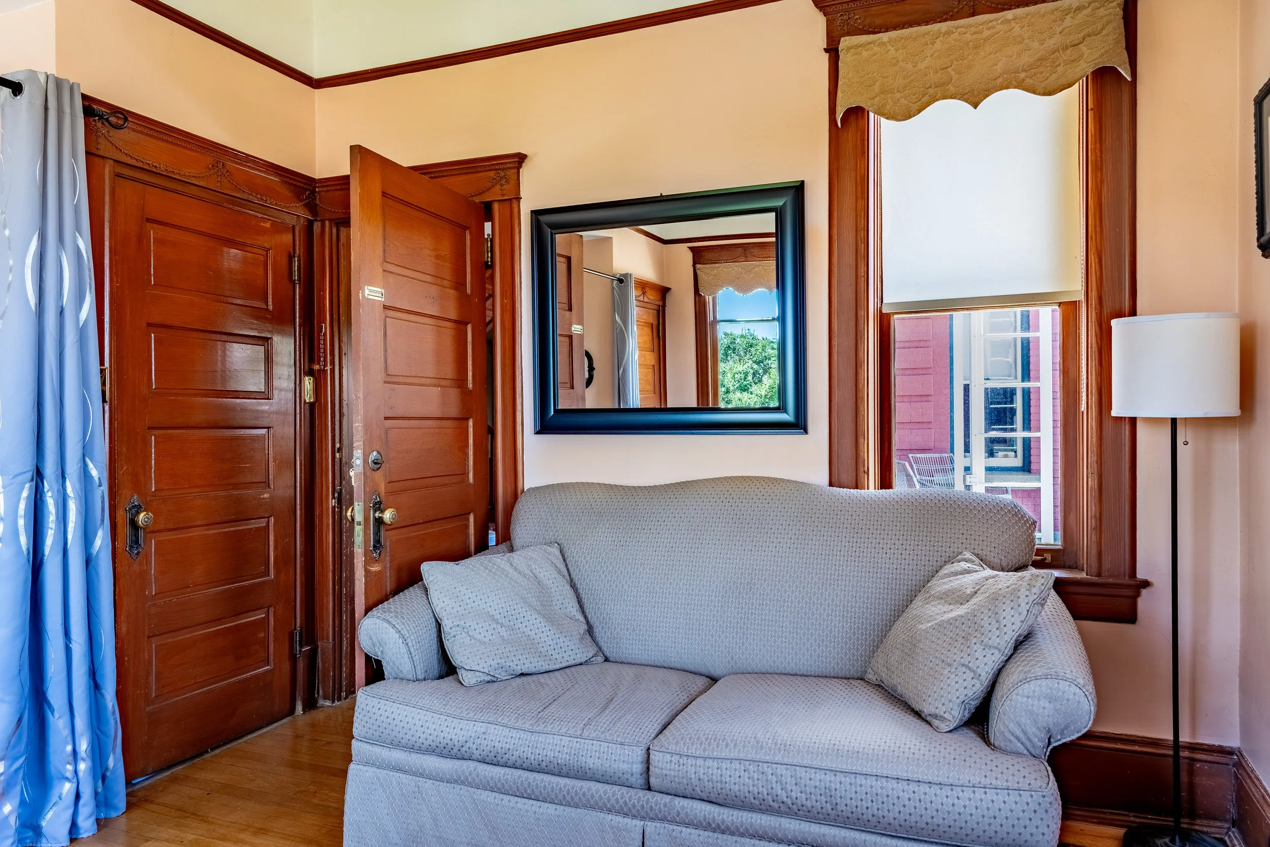 Living room with open wooden door, window with a view of a house and trees outside, a framed mirror on the wall, and a beige sofa with cushions.