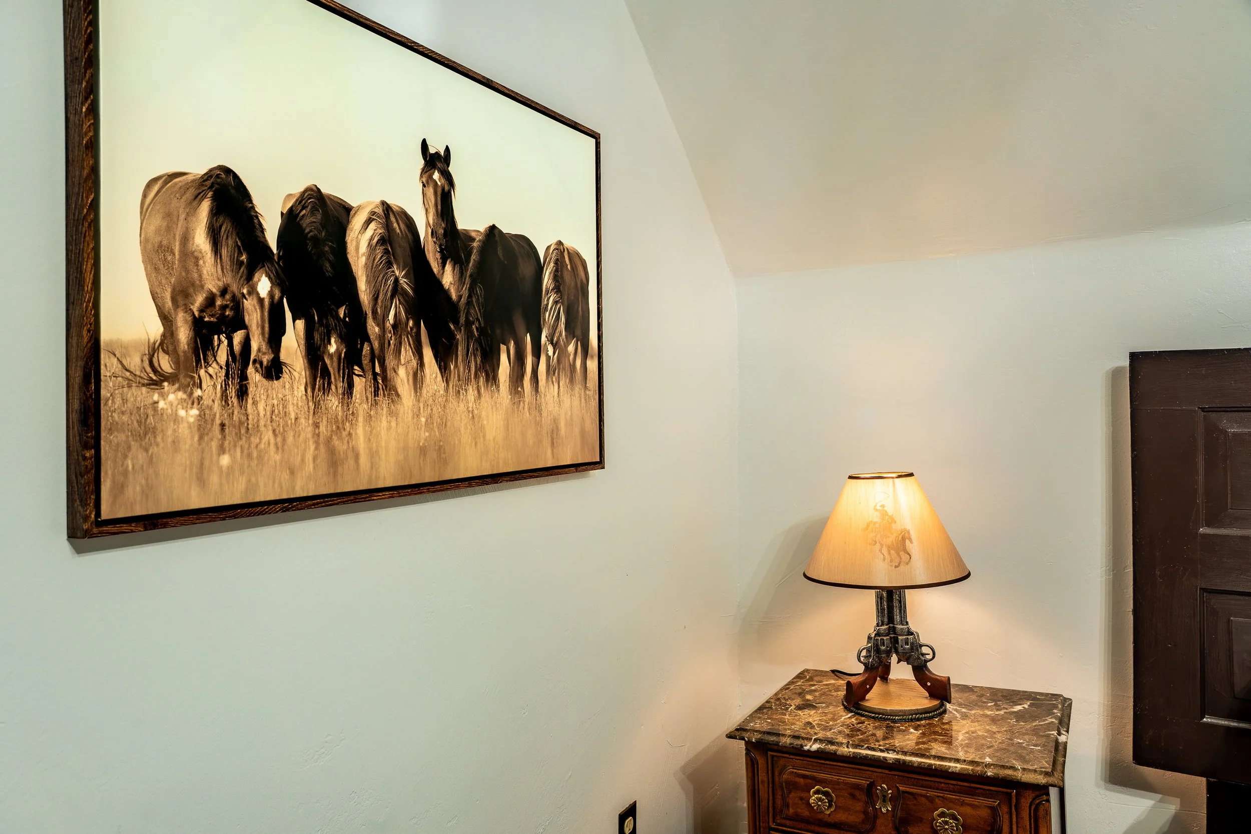 A framed photograph of seven horses in a field hangs on a light wall next to a table with a patterned marble top and a lamp with a beige shade, in a cozy room.