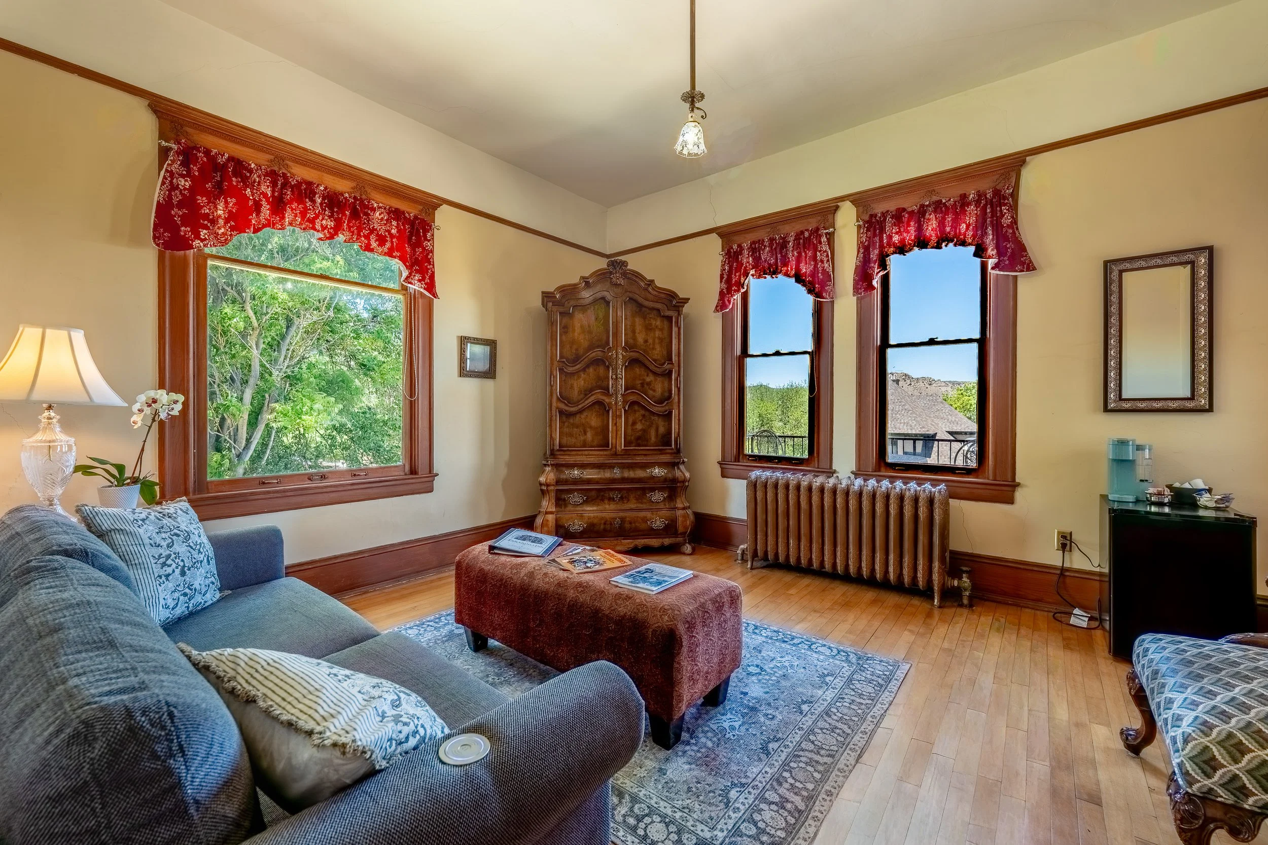 A living room with a blue sofa, pinkish-red ottoman, large windows with red floral valances, vintage wooden wardrobe, radiator, table lamp, mirror, and view of trees and houses outside.
