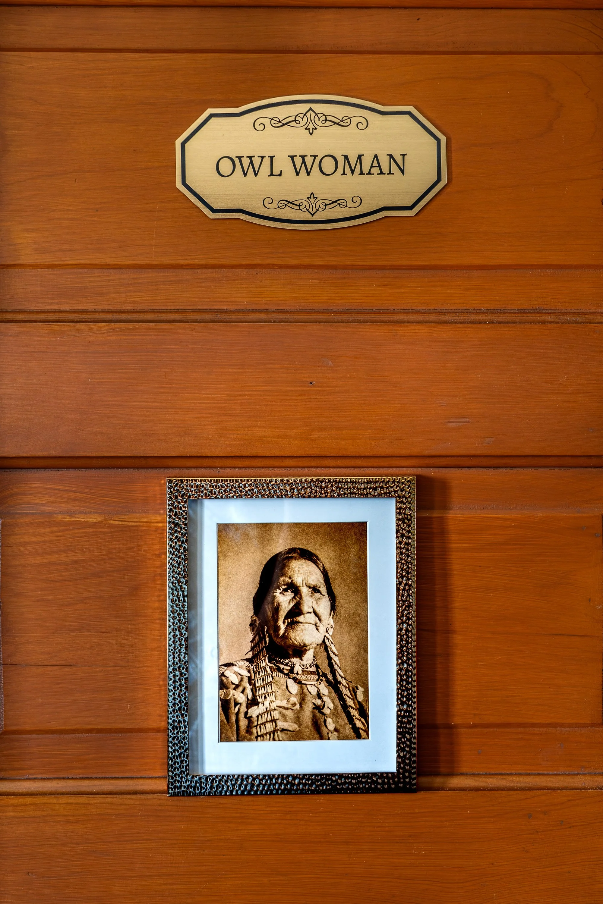 A wooden wall with a sign that reads 'OWL WOMAN' and a framed black-and-white portrait of an elderly Native American woman wearing traditional clothing and jewelry.