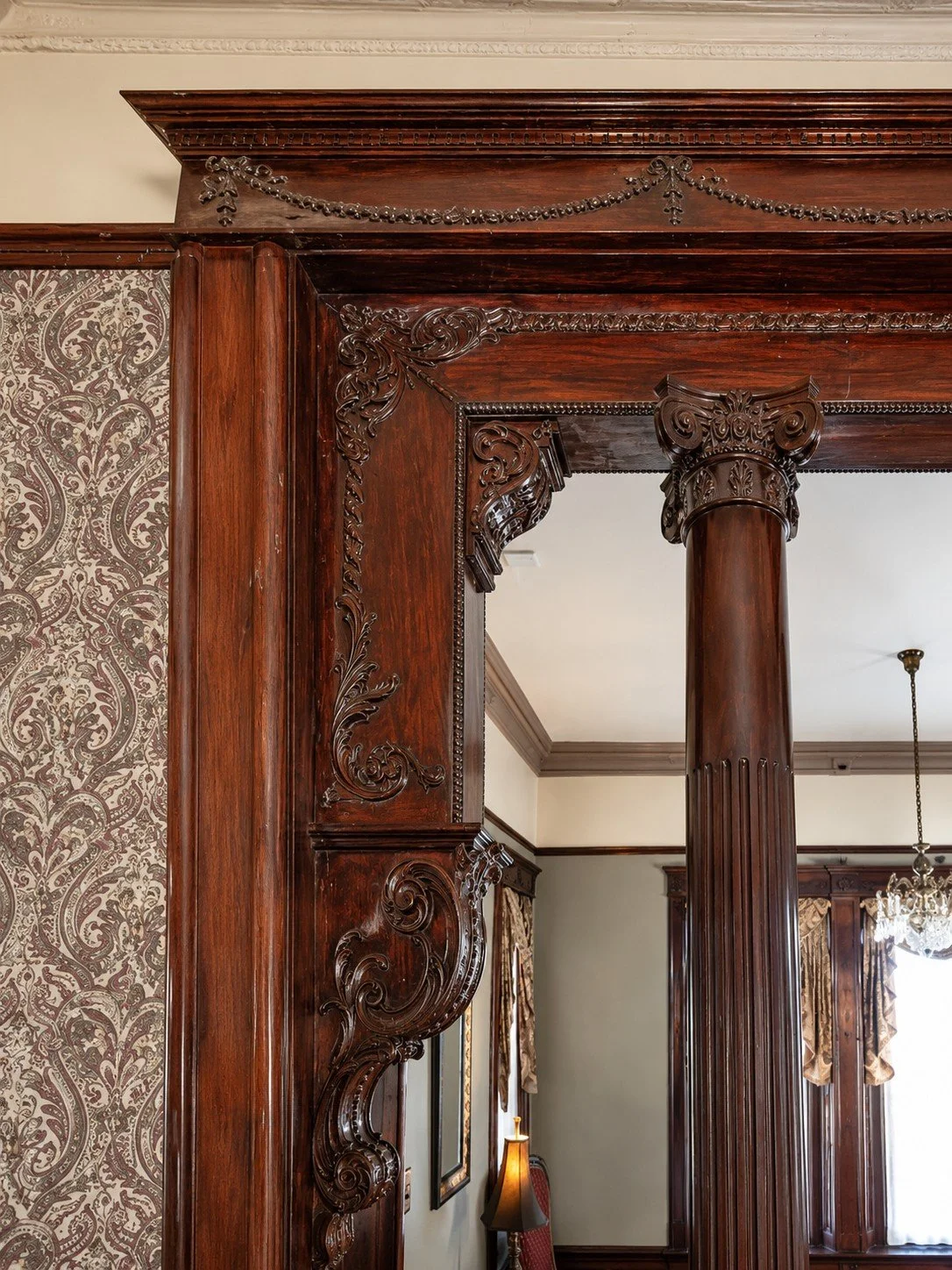 Close-up of ornate, dark wood decorative trim and a column in a room with vintage decor, including patterned wallpaper, a chandelier, and drapes.