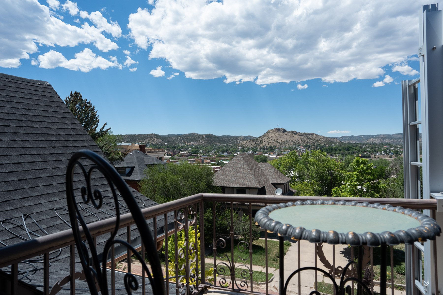 View of a neighborhood with rooftops and trees, mountains in the background, blue sky with scattered clouds, and a balcony with a metal railing, a round table, and a patio chair.