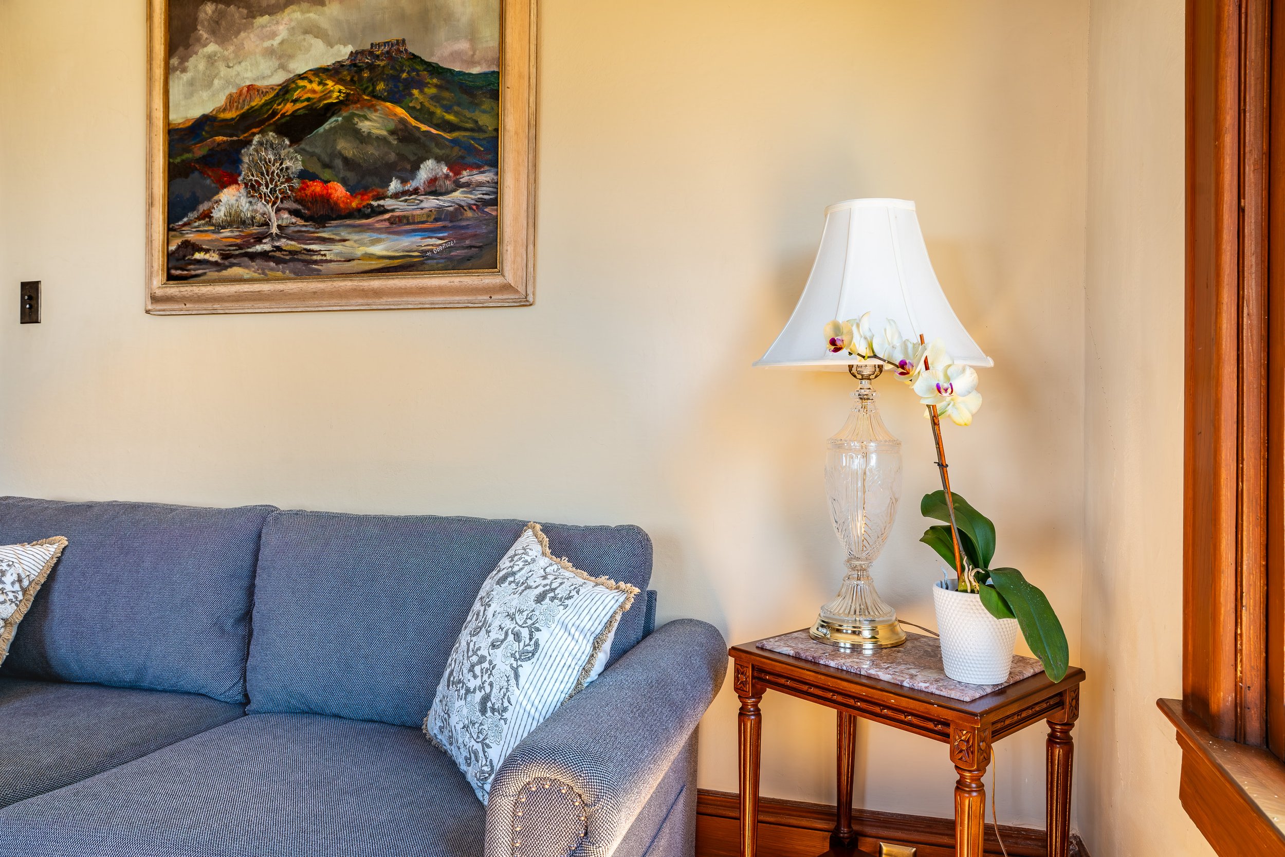 A living room corner with a gray sofa, decorative pillows, a framed landscape painting on the wall, a white lamp with a floral arrangement, and a wooden side table.