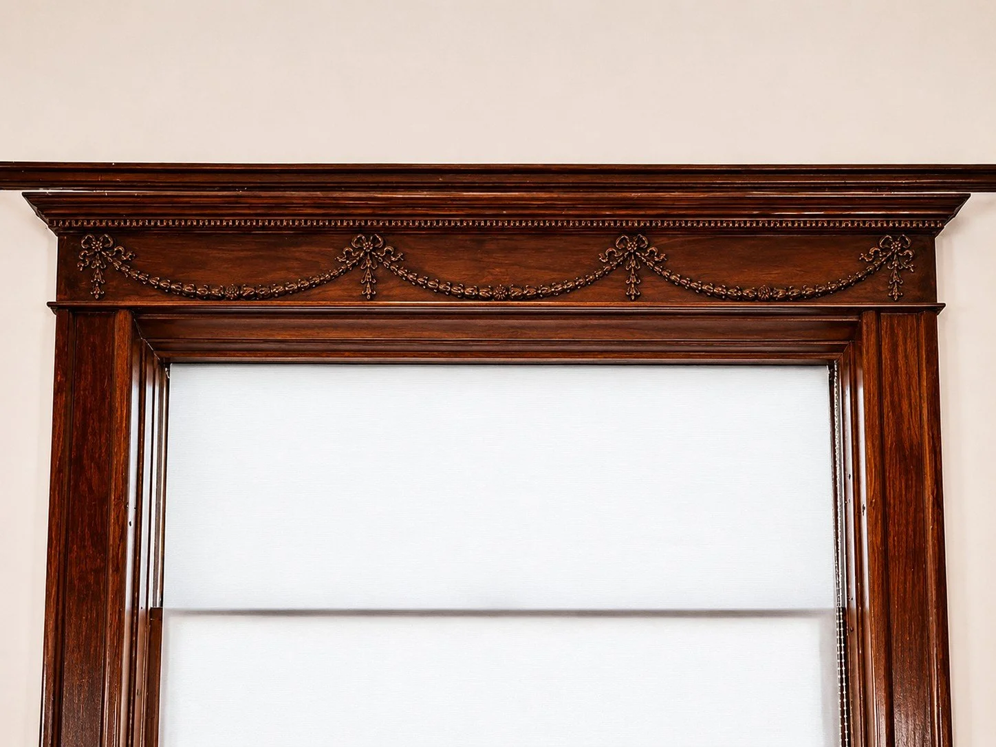 Close-up of a wooden window frame with intricate carved details and garland decoration on top, with a white window shade.