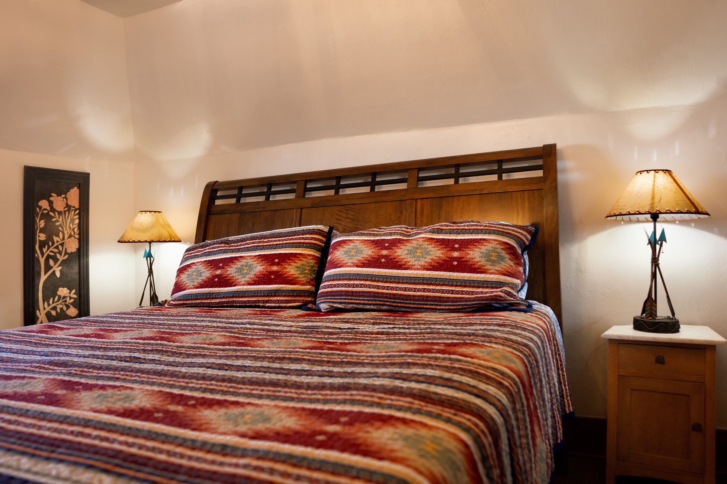 A cozy bedroom with two bedside lamps, wooden headboard, and patterned bedding, including pillows and a colorful quilt, under warm lighting.