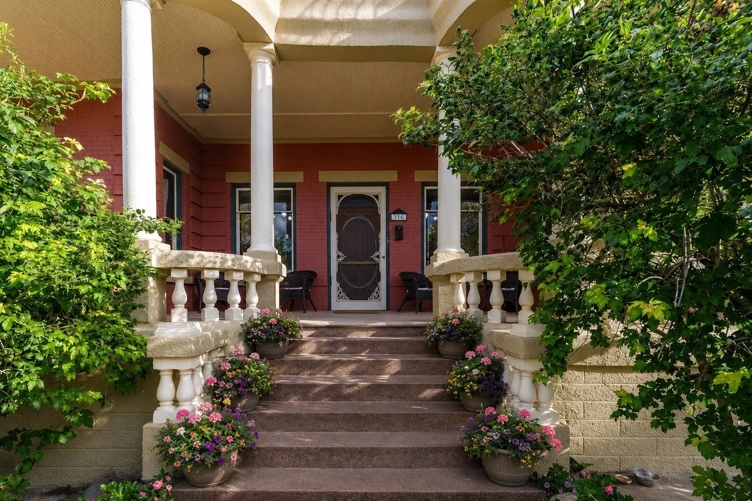 Front porch of a historic inn in Trinidad, CO, with red walls, white columns, and a black door, decorated with potted flowers on stairs and surrounded by green bushes.