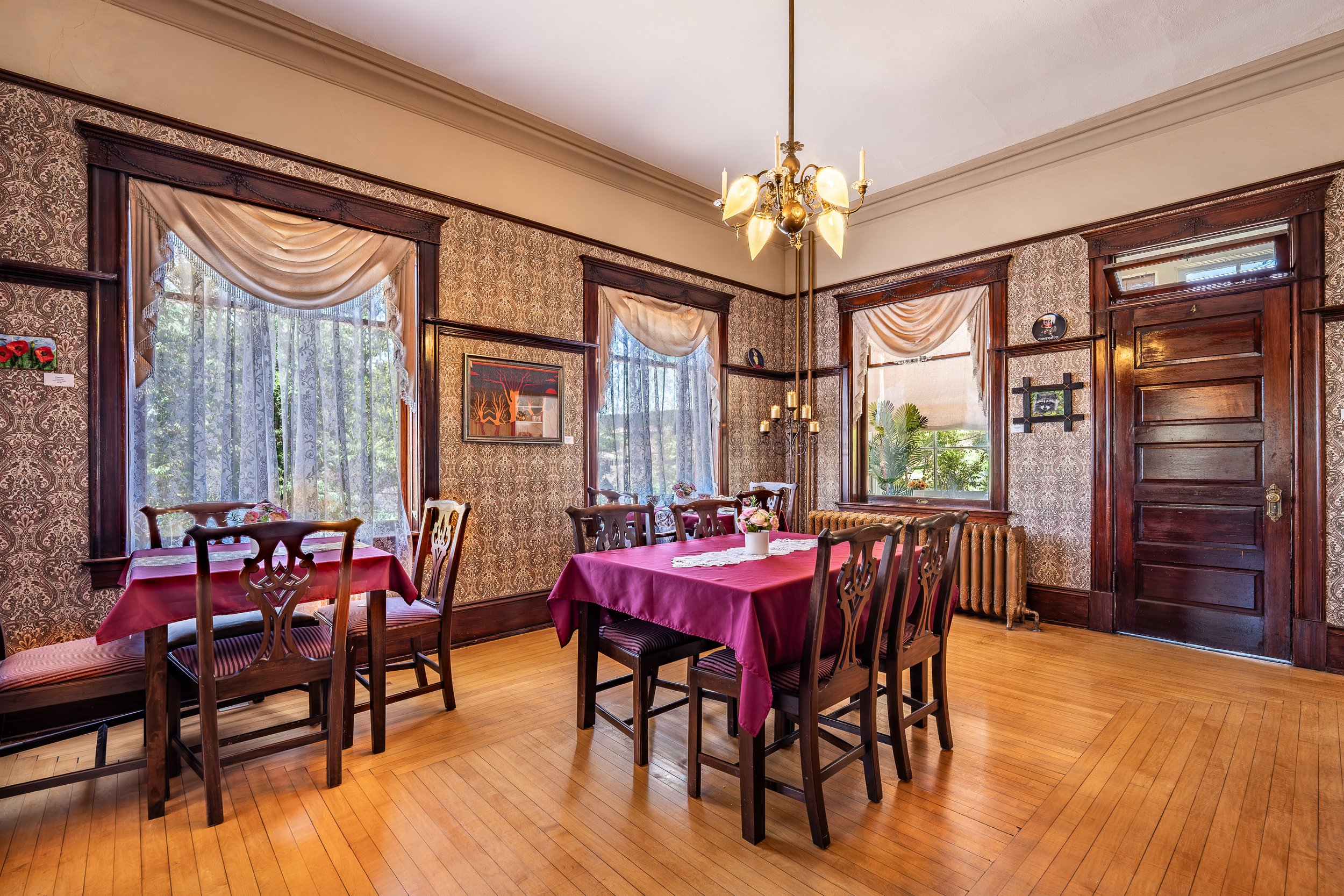 A cozy vintage dining room with two large windows dressed in sheer lace curtains and valances, floral wallpaper, a chandelier, multiple wooden chairs, and tables covered with pink tablecloths, set with small floral centerpieces, in a house with hardwood floors.