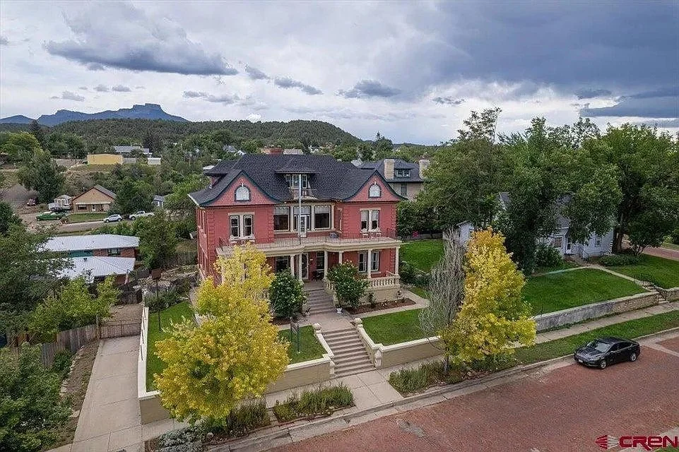 A large three-story Victorian-style house with a pink exterior and dark roof, surrounded by a landscaped yard with trees, sidewalk, and a driveway, located in a suburban neighborhood with other houses and greenery, under a cloudy sky.
