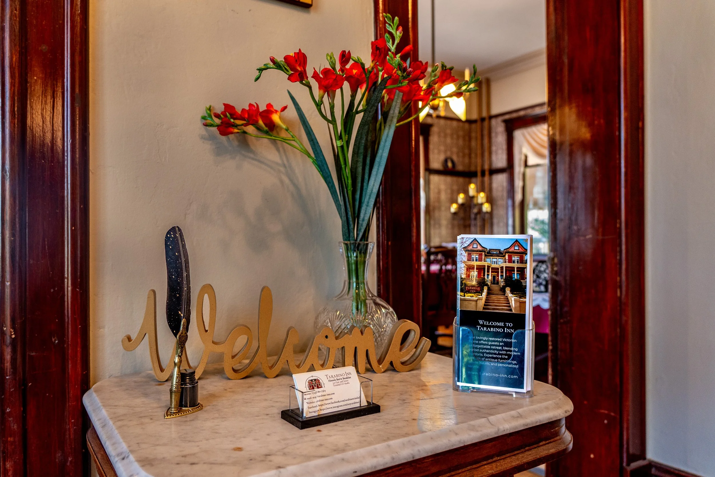 A marble table with a decorative sign. A clear glass vase with red flowers on the table. A wooden 'welcome' sign. A brochure or flyer with details about Tarabino Inn. The background shows wooden door frames and a glimpse into a living area with lit chandeliers.