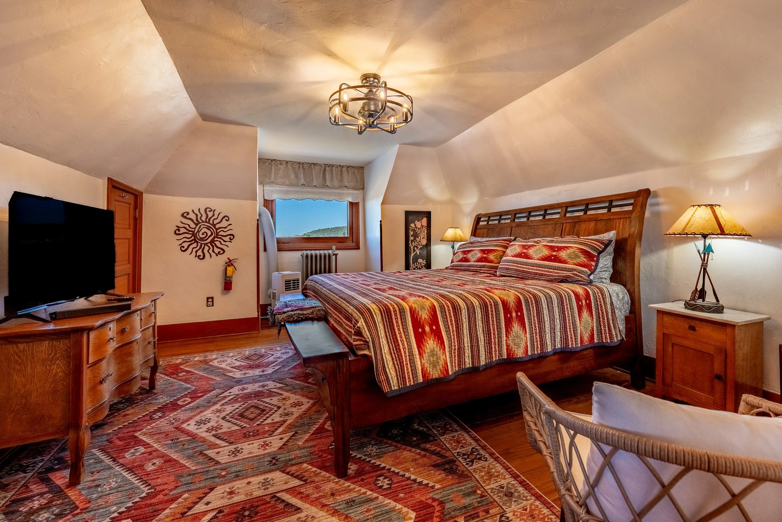 A cozy bedroom with sloped ceilings, featuring a large wooden bed with colorful patterned bedding, two bedside lamps, a window with a scenic view, and a ceiling light fixture.