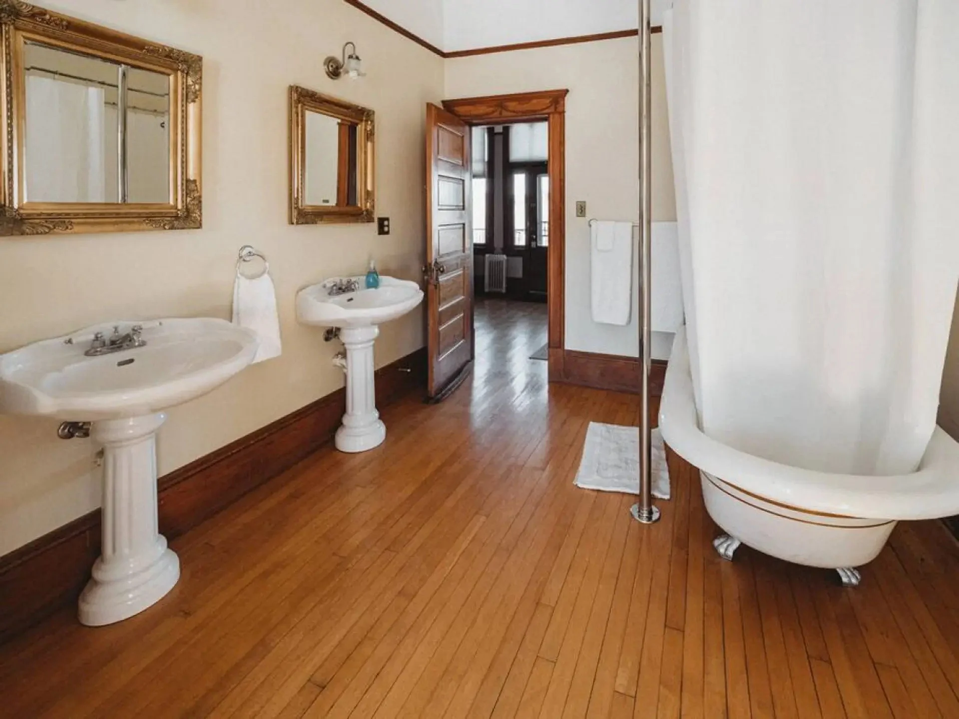 A vintage-style bathroom with hardwood floors, two pedestal sinks, framed mirrors, a wooden door leading to a sunlit room, and a white clawfoot bathtub with a shower curtain and a chrome shower rod.