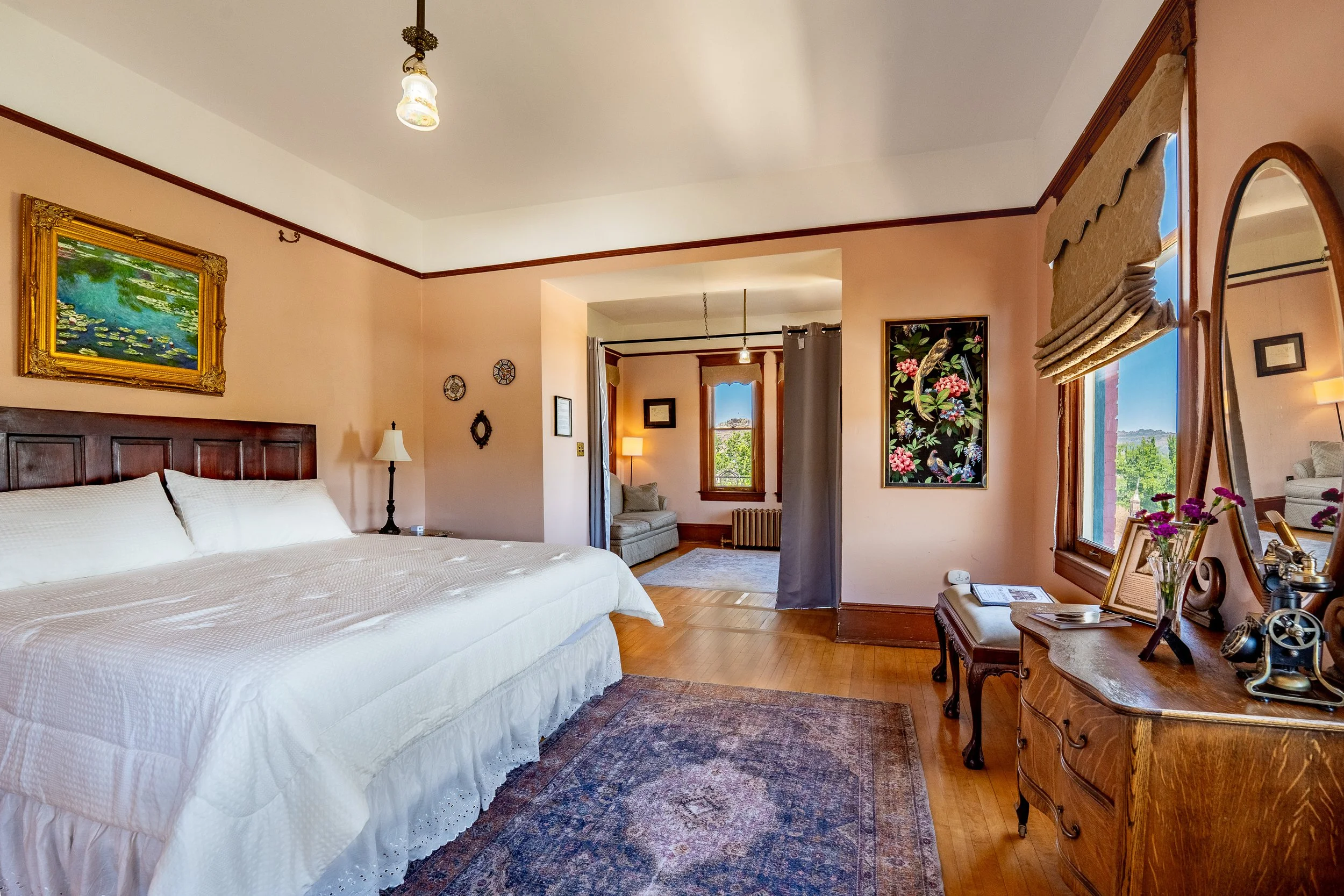 A cozy bedroom with a large bed covered in white bedding, artwork on the walls, a window with beige valances, a wooden dresser with decorative items, and a sitting area by a window in the background.