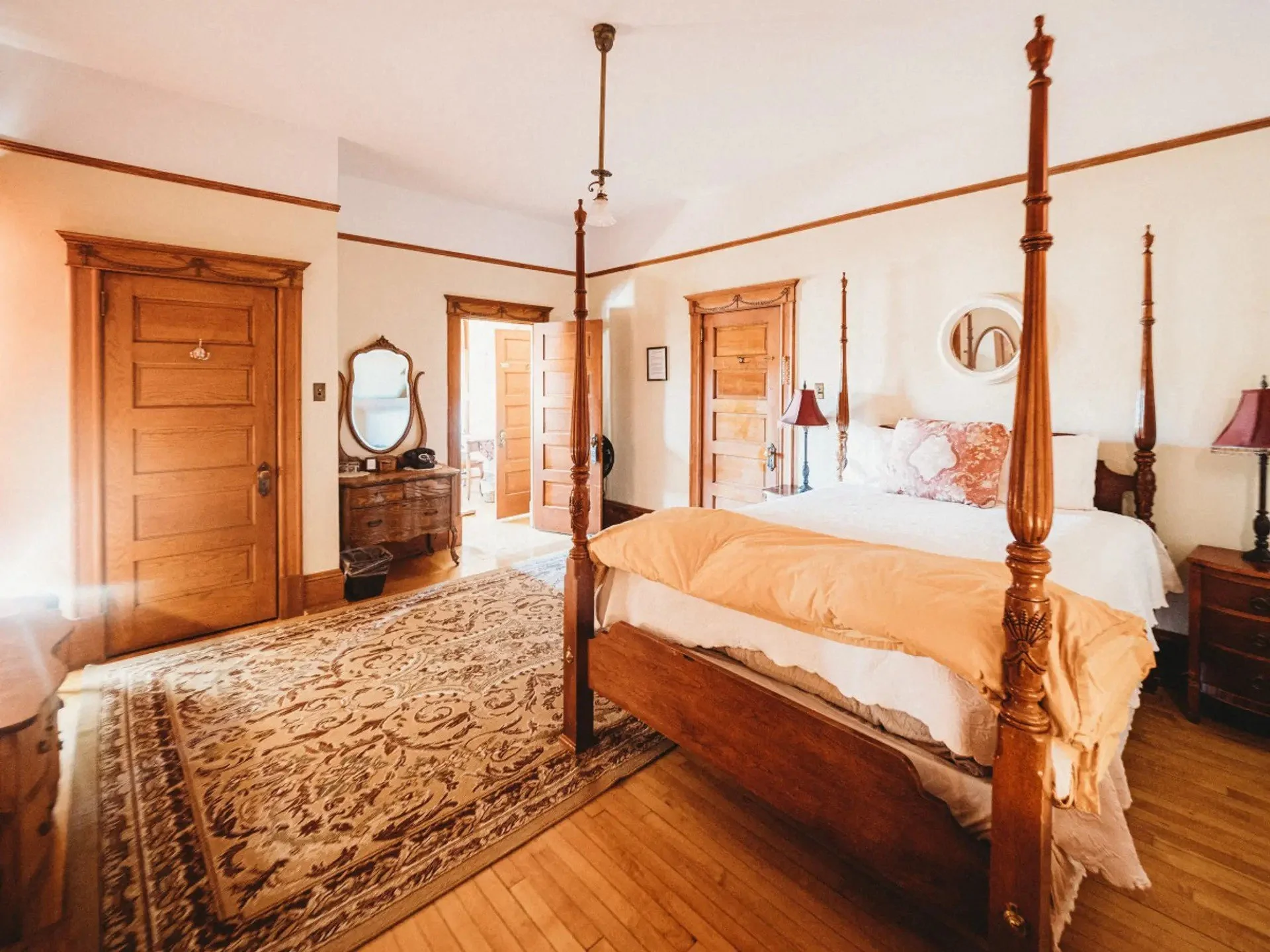Bedroom with wooden furniture, four-poster bed, bedside tables, dresser with mirror, wall mirror, lamps, wooden doors, and a patterned area rug.