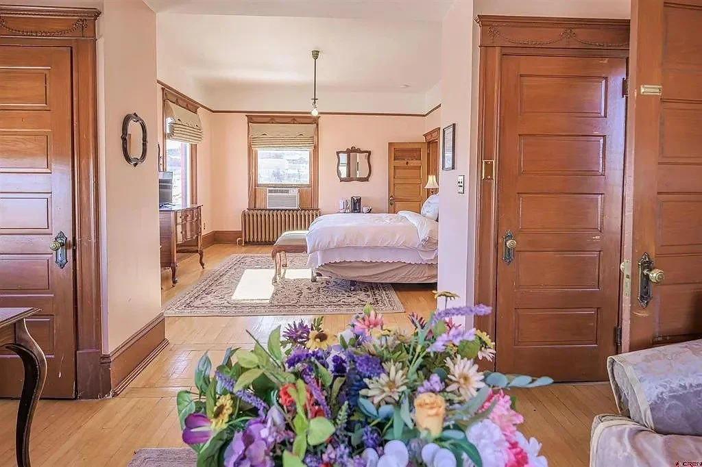 A cozy bedroom with wooden doors and trim, a bed with white bedding, an area rug, a window with blinds, and a bouquet of colorful flowers in the foreground.