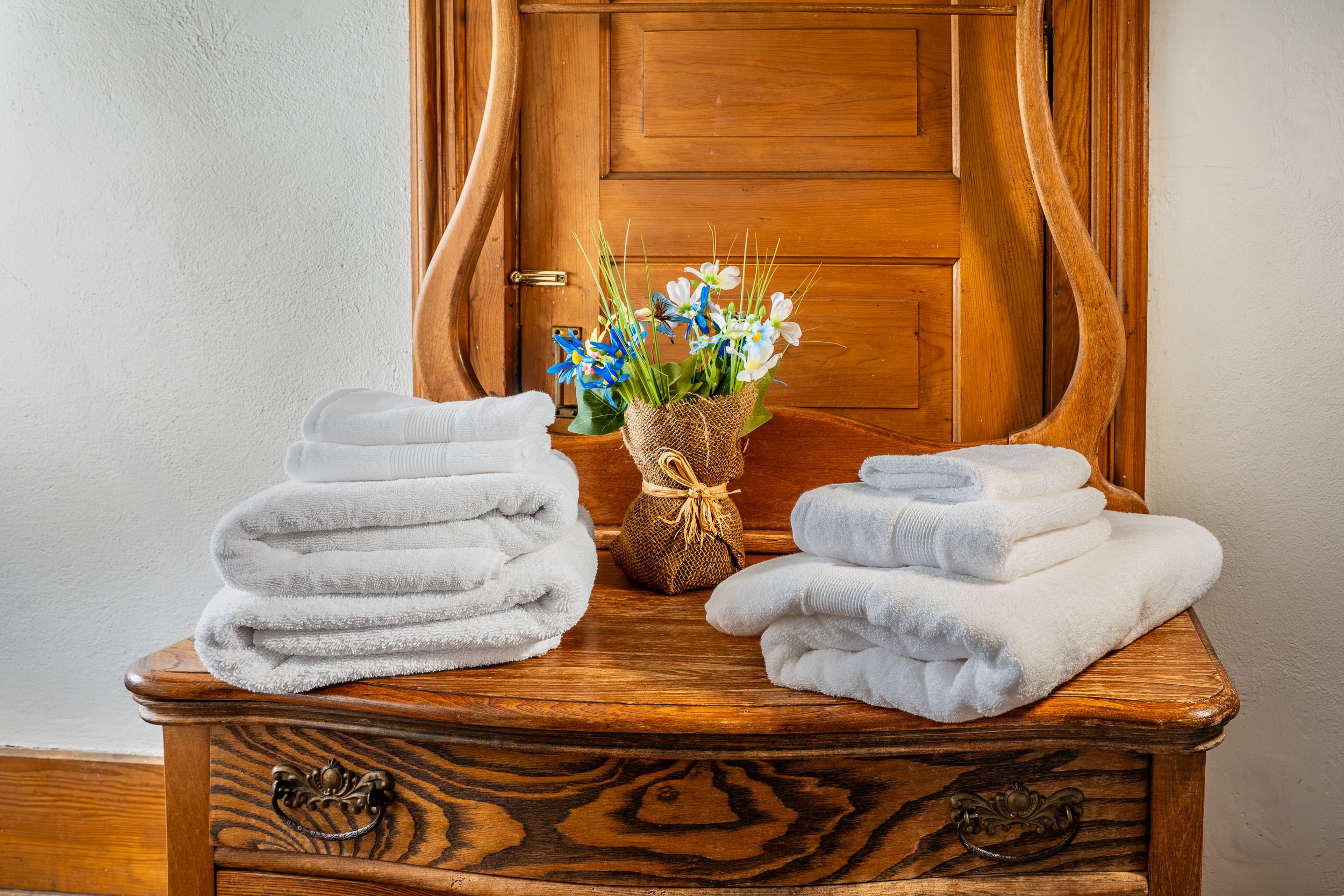 A wooden dresser with a mirror, topped with folded white towels, a bouquet of white and blue flowers in a burlap-wrapped vase, set against a white wall.