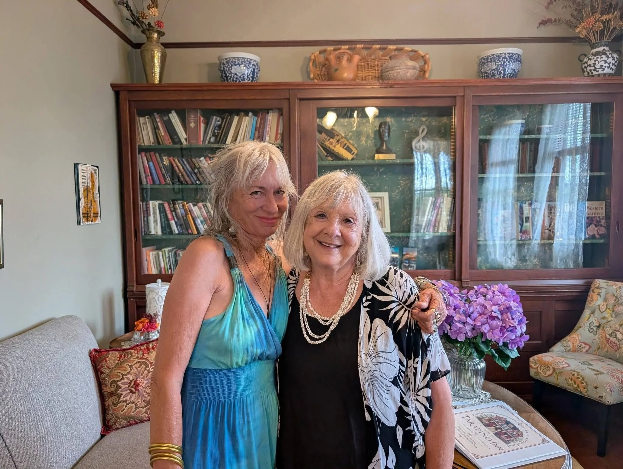 Two women with white hair smiling while standing behind a table with purple flowers in a glass vase. They are inside a room with a large wooden bookcase and decorative vases and artwork in the background.