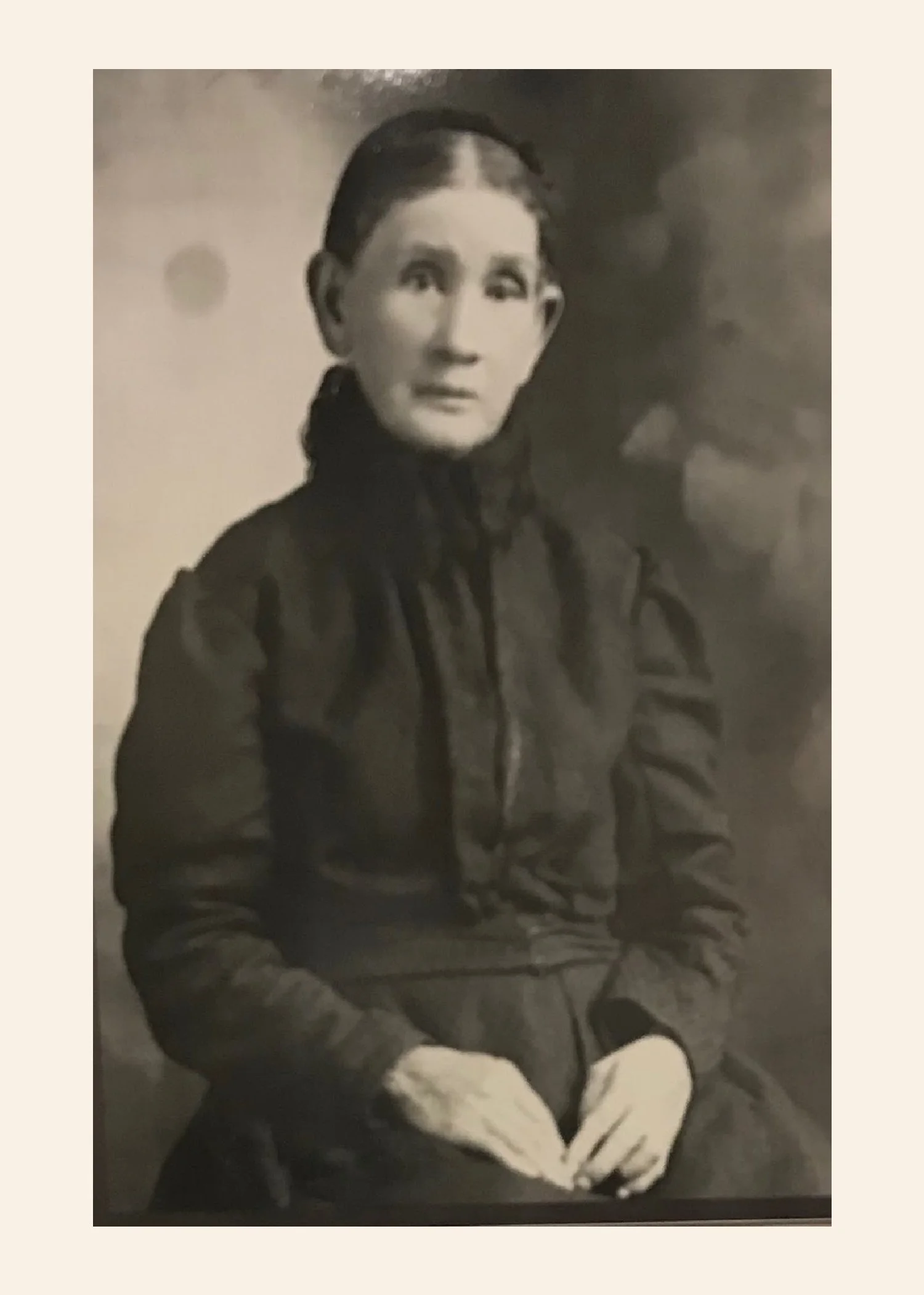 Black and white photo of a young woman with dark hair pulled back, wearing a jacket, sitting with her hands clasped in her lap, looking slightly to her left.