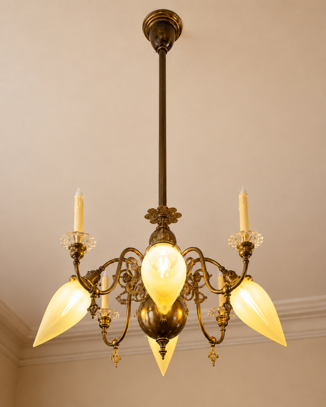 An ornate brass chandelier with five yellowish glass light fixtures and four candles hanging from a ceiling.