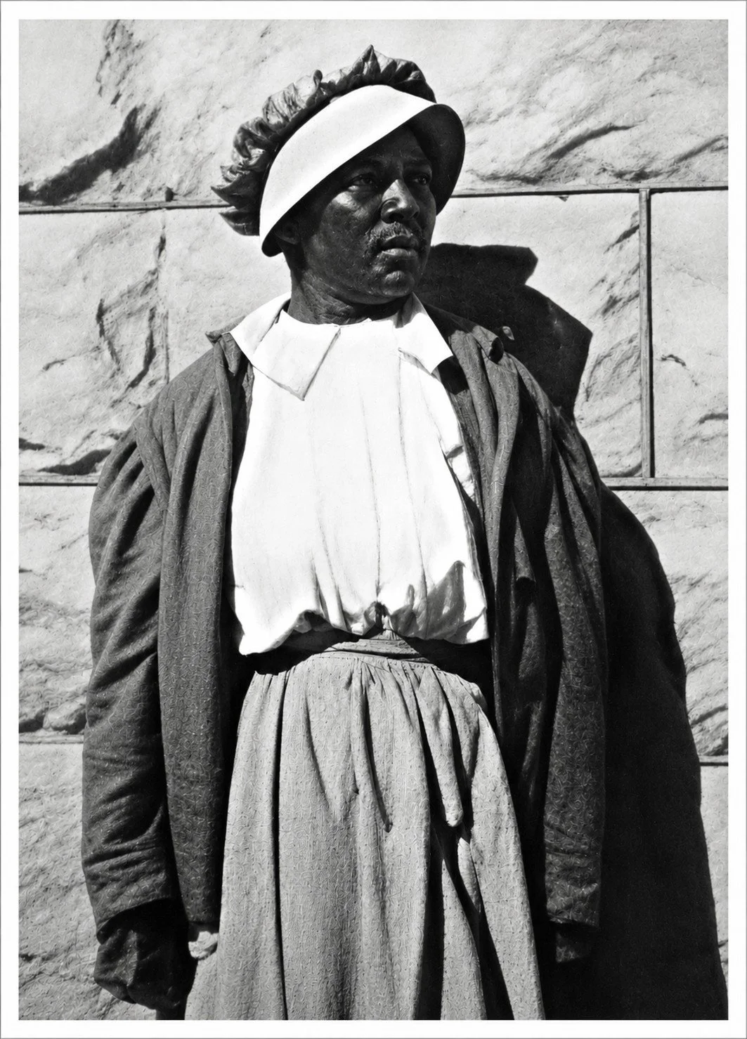 A woman dressed in historical clothing, including a hat, blouse, and skirt, standing in front of a marble wall.