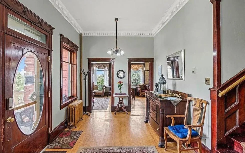 Foyer of a historic-style home with wooden floor, antique furniture, and framed decor, leading into a living room with large windows and natural light.