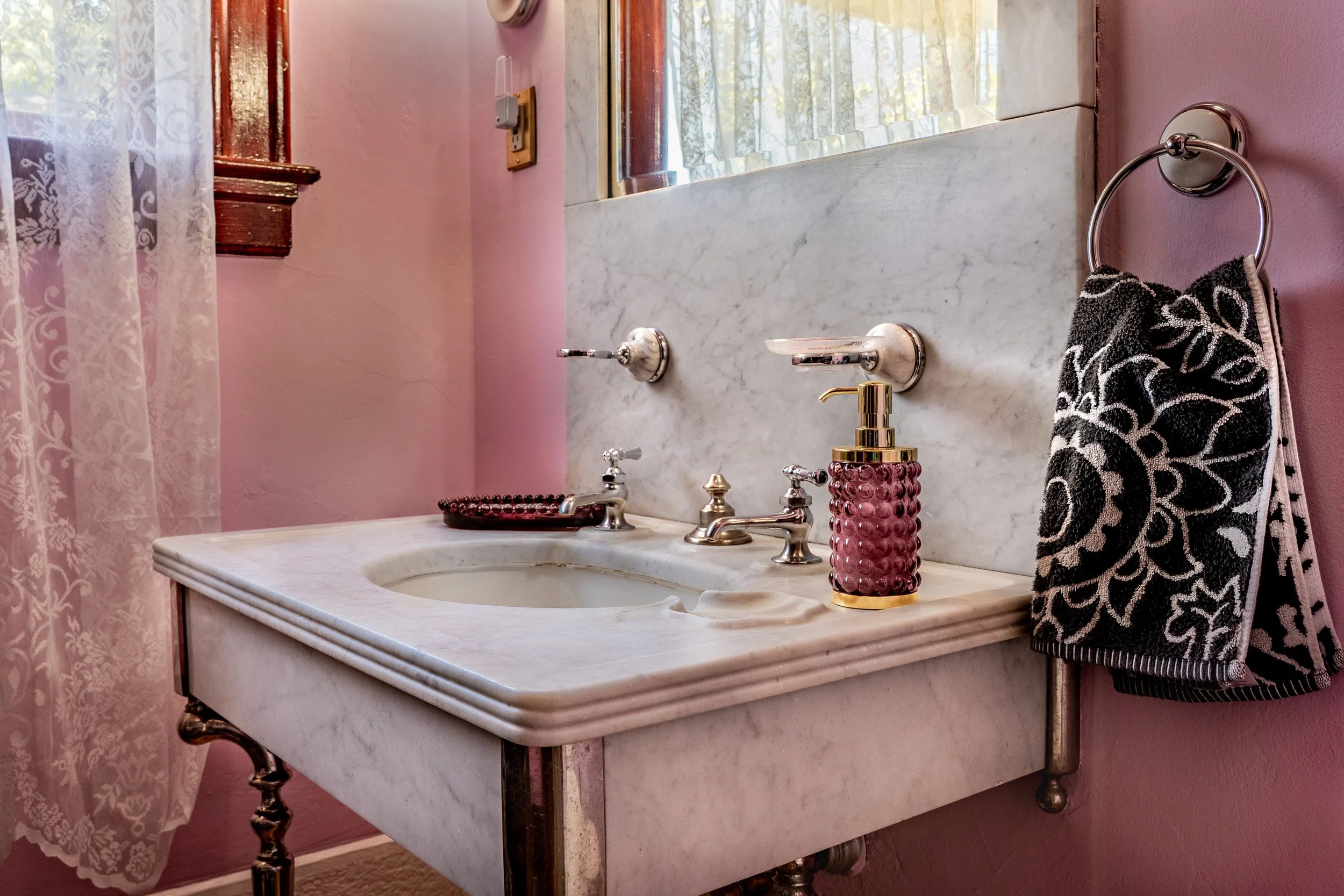 A vintage bathroom sink with a marble countertop, chrome fixtures, a pink textured soap dispenser, and a black-and-white patterned hand towel hanging on a chrome ring. A mirror and window are reflected in the background, with pink walls and a lace curtain.
